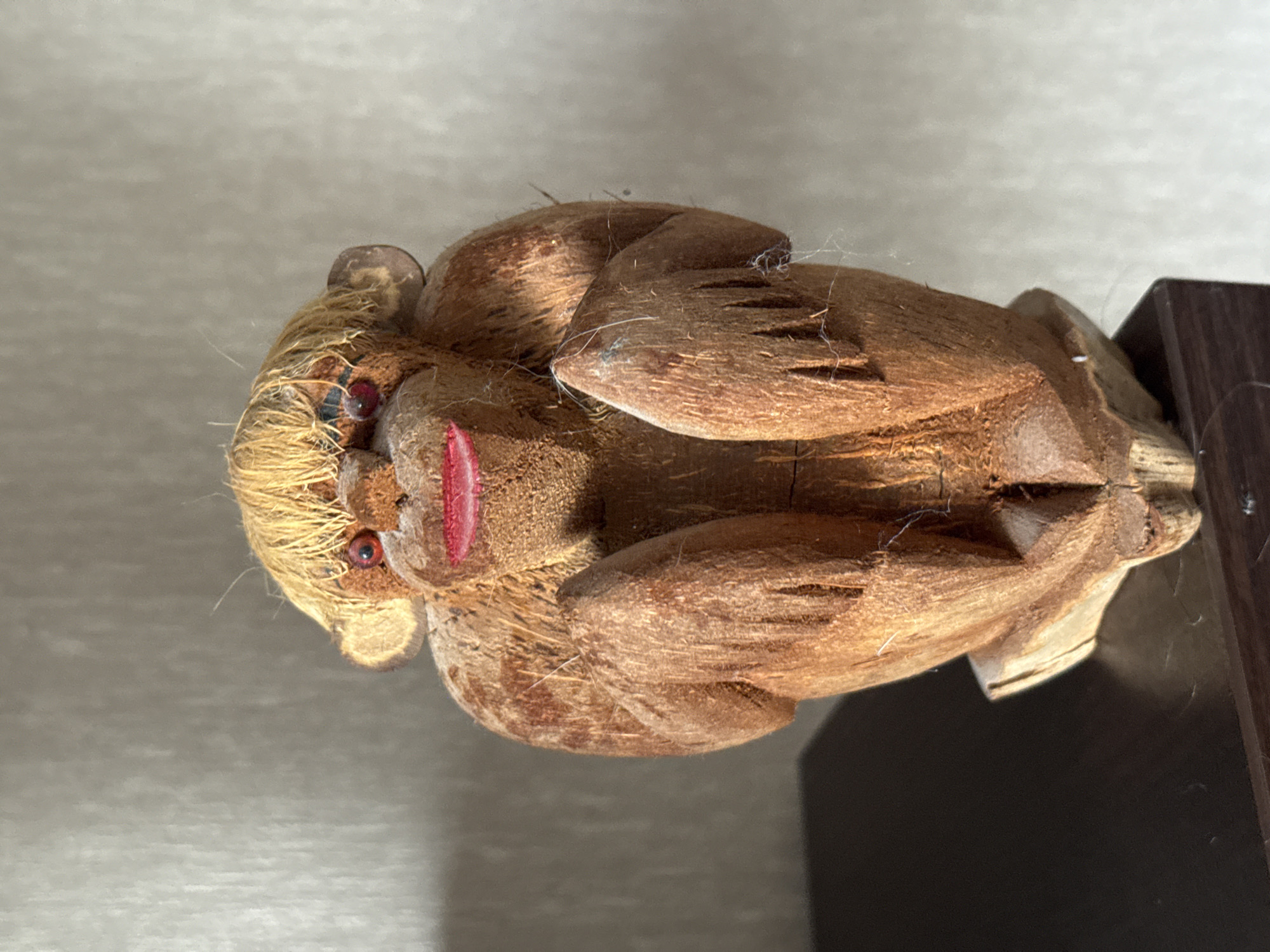 A wooden monkey figurine, carved from coconut shell material, is the subject of the image. The figurine has blonde hair made of straw-like material, red eyes, and bright red lips. It is sitting with its arms bent and resting in front of its body. The monkey is positioned on a dark, flat surface that appears to be a table or shelf. The background is a textured, light-colored wall, possibly with subtle vertical lines. The lighting suggests a natural light source coming from the side, casting shadows on the left side of the figurine and the surface it rests on. The overall scene is a close-up, frontal view of the handcrafted object. The location is not specified within the image or its context.