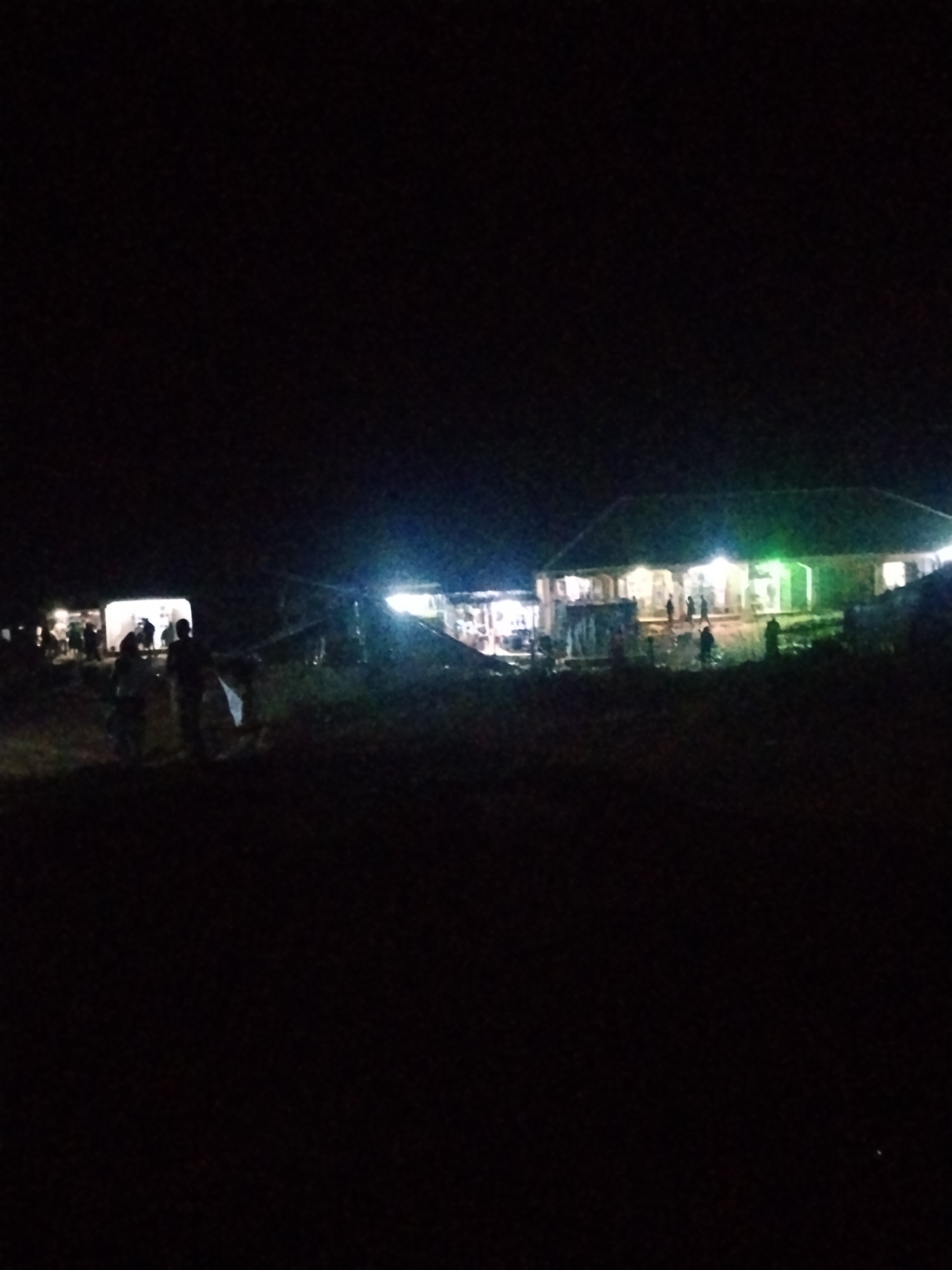 The image captures an outdoor gathering at night in Durumi, Nigeria. Several structures are illuminated, suggesting a communal event or gathering. In the foreground, silhouettes of people are visible, walking or standing. Further back, a larger building, possibly a community hall or a long house, is brightly lit, with several individuals gathered around its entrance. To the left, a smaller, brightly lit tent or temporary structure is also visible, with people congregating nearby. The scene appears to be on uneven ground, with some vegetation or debris scattered around. The time of day is clearly night, indicated by the darkness of the sky and the reliance on artificial lighting. The lighting from the structures casts a glow on the surrounding area, creating a lively atmosphere. The overall impression is of a community event taking place after dark. There is no visible text in the image.