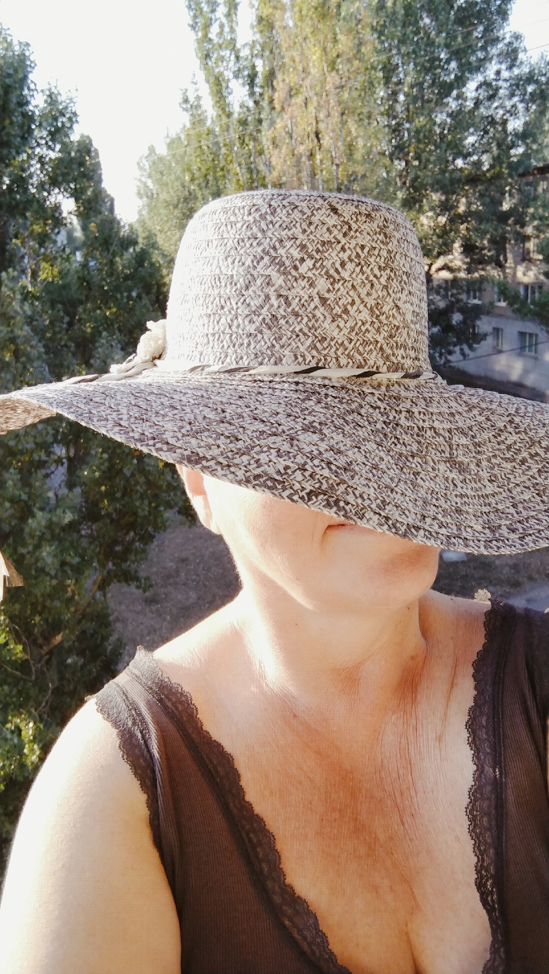 A person wearing a wide-brimmed straw hat and a dark lace-trimmed top is shown from the chest up. The hat obscures most of the person's face. The background consists of trees and buildings, suggesting an outdoor urban or suburban setting. The lighting indicates it is daytime. The image was taken in Horishni Plavni, Ukraine.