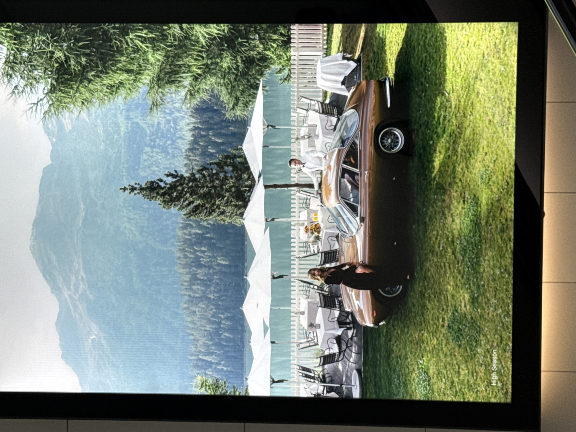 The image depicts a scene at a lakeside resort with a mountain backdrop. In the foreground, a vintage brown convertible car is parked on a manicured lawn. A woman in a black dress is leaning against the car. Behind the car, there are several white umbrellas shading outdoor dining tables, a white picket fence, and a body of water, likely a lake, with a forested hillside beyond. Further in the background, a mountain range is visible under a hazy sky. The overall impression is one of luxury and leisure. The text "High Season" is visible in the bottom left corner of the image, suggesting a promotional or artistic context. The scene is not located in Sesto San Giovanni, Italy, as this is an outdoor scenic view typical of alpine regions.