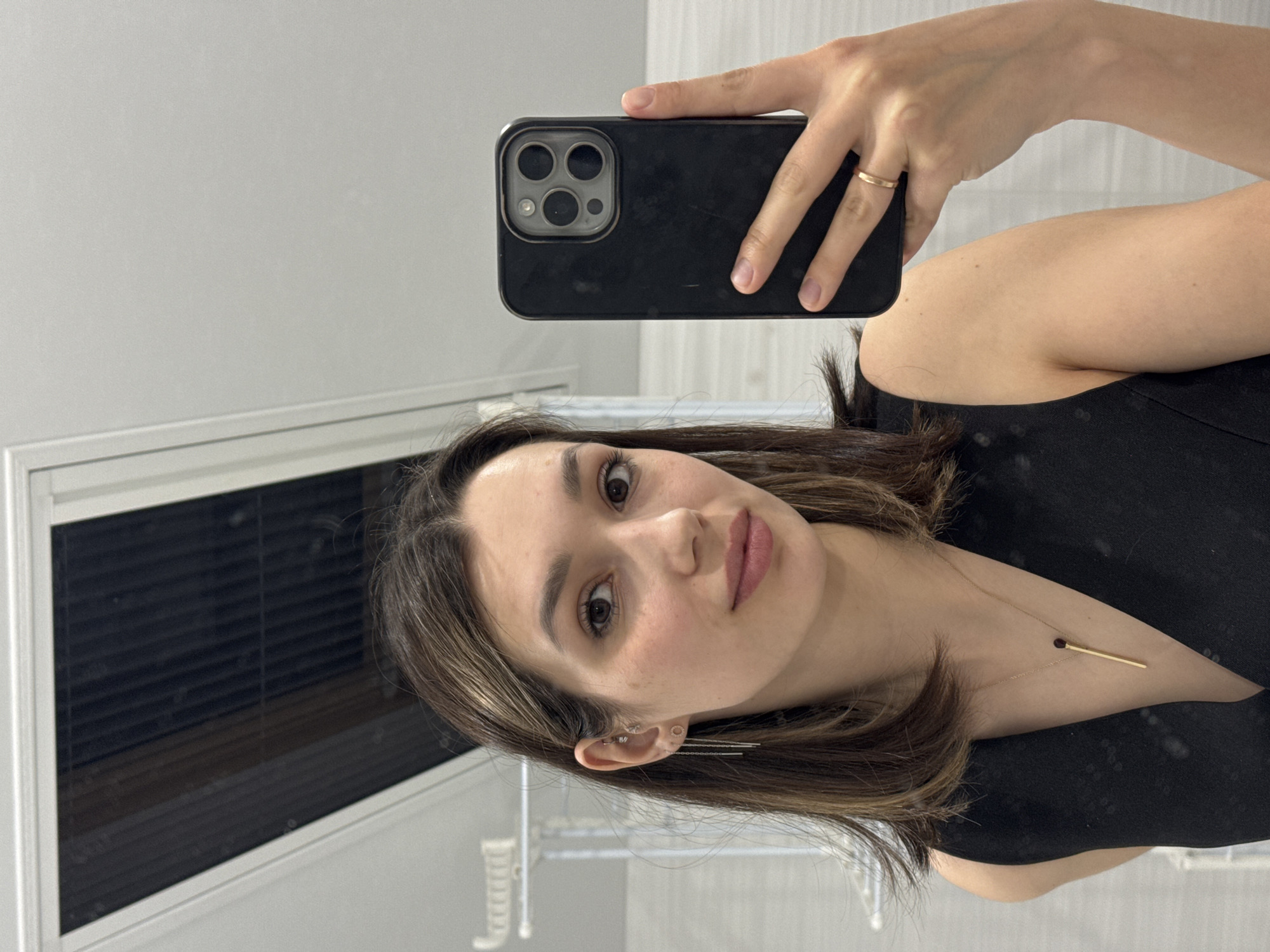 A person is taking a selfie with a black phone in front of a reflective surface. The person has shoulder-length brown hair with blonde highlights, and is wearing a black sleeveless top and gold jewelry, including a ring and necklace with a matchstick pendant. The background appears to be a room with light grey walls and a window with dark blinds. The city and precise location are unknown.