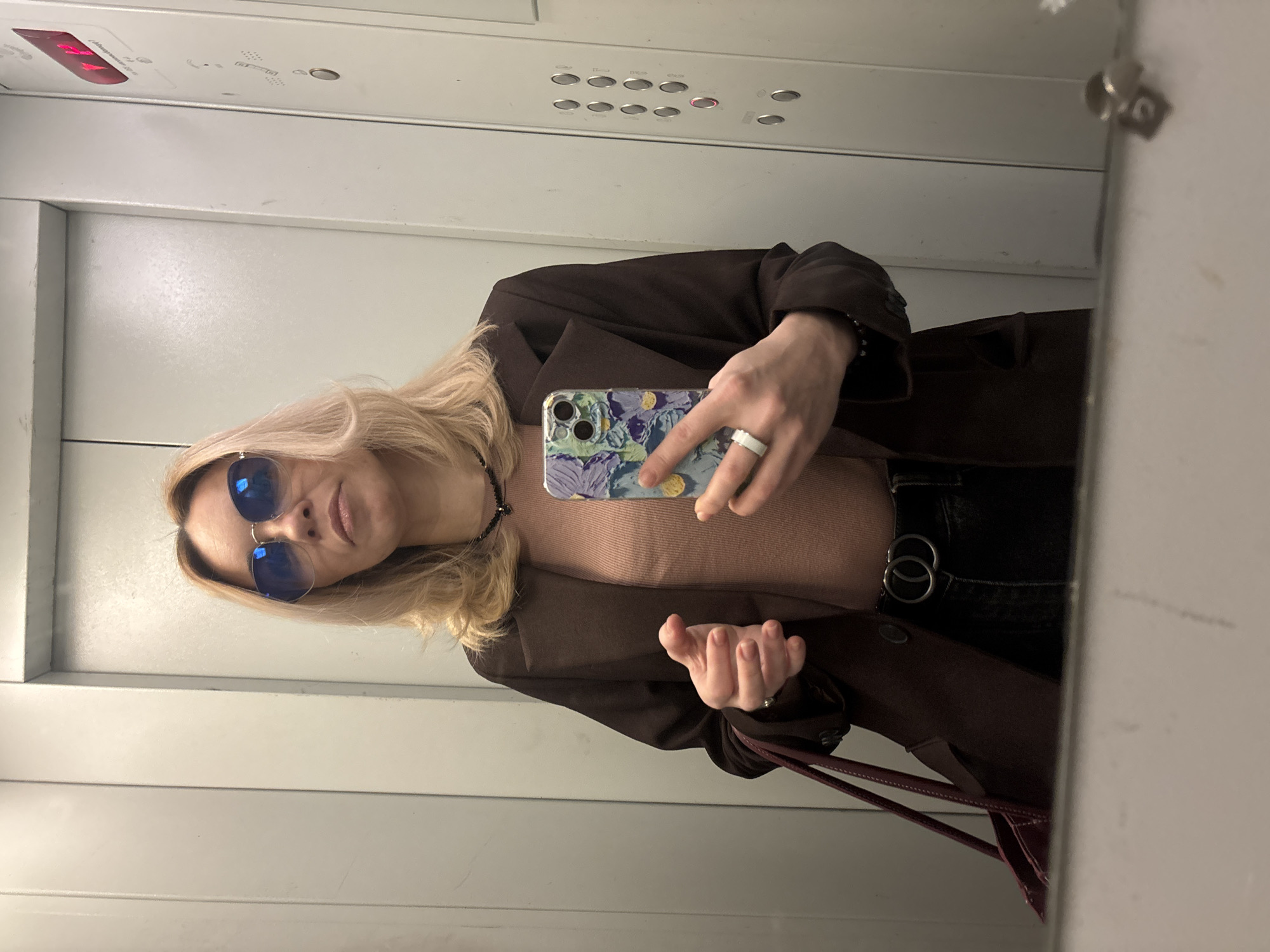 The image is a selfie of a blonde woman wearing sunglasses and a brown jacket in an elevator. She is holding a phone with a patterned case. The elevator has a control panel and a light. The woman is also wearing a necklace, a light pink shirt, and a black belt. The location is likely Vsevolozhsk, Russia, as requested, but there is no specific evidence of this in the image.