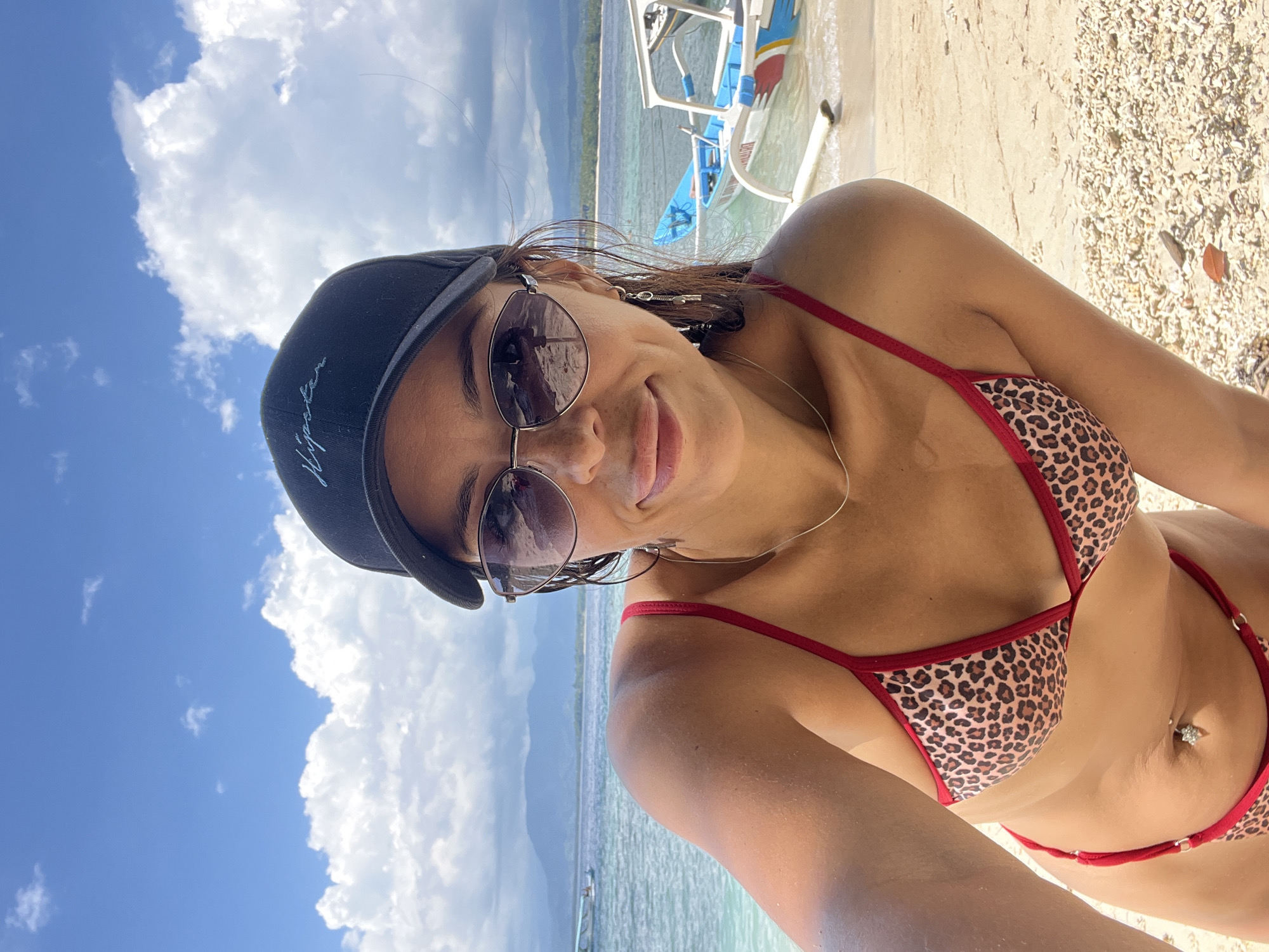 A woman is posing for a selfie on a beach in Unknown, Indonesia. She is wearing a black baseball cap and sunglasses, and a brown and red leopard print bikini. Behind her is a blue and white boat, a blue body of water, and the sand on the beach. The sky is visible above, with white clouds.