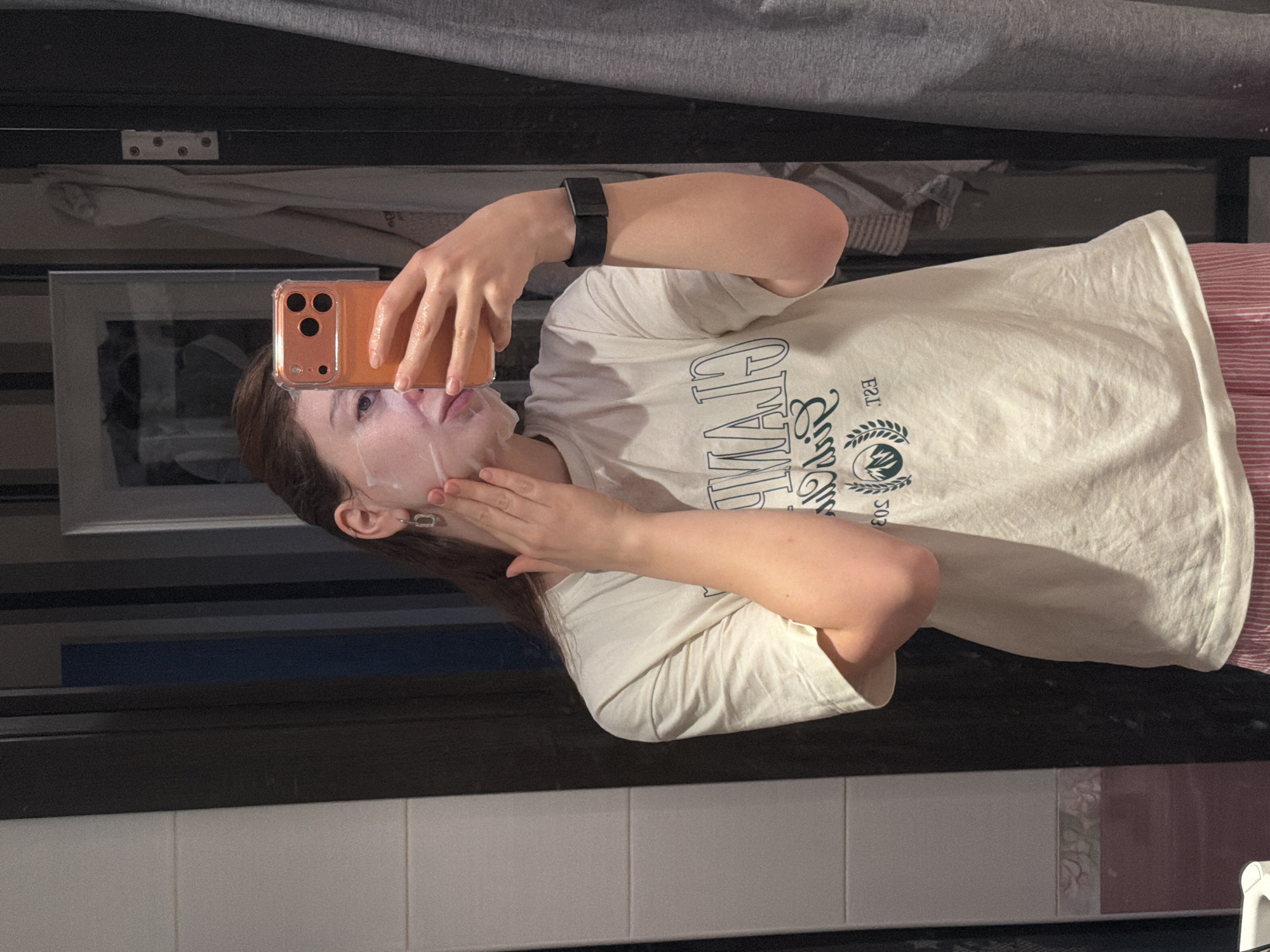 An indoor mirror selfie shows a person in Khimki, Russia, applying a face mask. The individual, with long brown hair, wears a light-colored (cream or off-white) t-shirt that displays mirrored text, likely reading "CHAMPION" (or similar) with "EST." and a date (e.g., "1993" or "503") and a small emblem such as a wreath or mountain design. A dark-colored smart watch is visible on their left wrist.
The person holds an orange-cased smartphone (appearing to be an iPhone model) in their right hand, using it to take the photograph. Their left hand gently presses the white face mask onto their face. The background, seen as a reflection in the mirror, features a dark rectangular frame, a grey curtain or fabric, and a wall partially covered with white tiles. A decorative strip of pink/maroon tiles with a subtle floral pattern is visible towards the bottom right. Light-colored, possibly striped, pajama pants can be seen at the edge of the frame. The scene is illuminated by indoor artificial lighting, suggesting the time is likely evening or night. The person's expression is neutral, focused on the self-care routine and taking the picture.