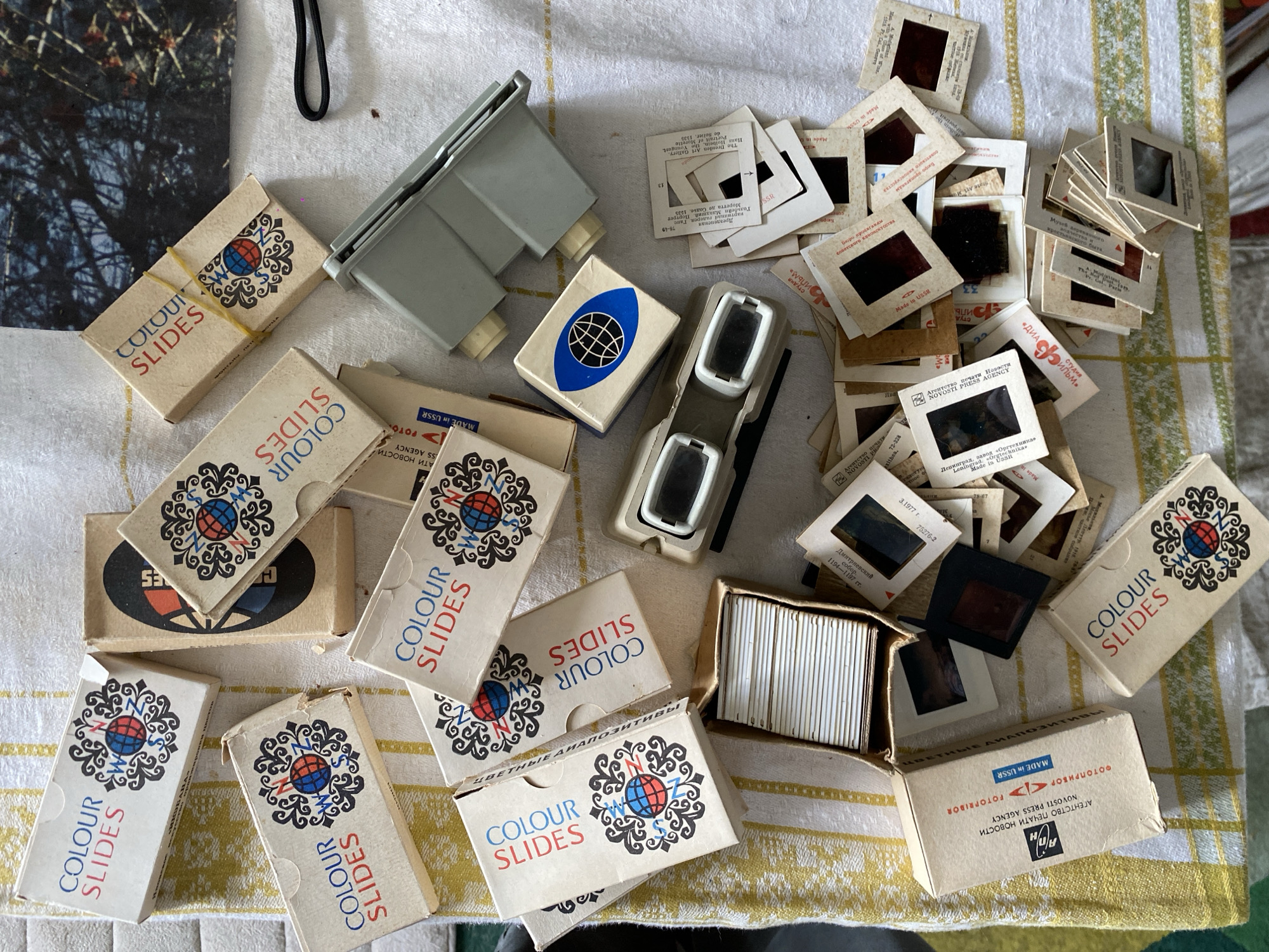 An overhead shot displays a collection of vintage photographic slides and associated equipment laid out on a patterned, yellow and white striped tablecloth.
The main subjects are numerous cardboard-mounted photographic slides, both loose and partially organized. Many are housed in small, aged cardboard boxes prominently labeled "COLOUR SLIDES" in English, featuring a distinctive red and blue globe logo with compass points. Several boxes also display Cyrillic text, including "ЦВЕТНЫЕ ДИАПОЗИТИВЫ" (Colour Diapositives).
Also visible are two small, handheld slide viewers—one gray, one white and gray—designed for individual slide inspection. A larger, light gray device, possibly a slide magazine or part of a projector, is situated near the top center. A single, partially visible photograph depicting tree branches against a dark background is located in the upper left corner. A black strap, perhaps from a camera or bag, can be seen at the top.
The arrangement suggests an activity of sorting, reviewing, or organizing a historical collection of photographs.
Notable details include the pervasive "MADE IN USSR" text on many slide mounts and boxes, clearly indicating their origin from the Soviet Union. Other visible text includes "NOVOSTI PRESS AGENCY" (АГЕНТСТВО ПЕЧАТИ НОВОСТИ), "ФОТОПРИБОР" (FOTOPRIBOR), and specific descriptions of slide content in both Cyrillic and English, such as "Дмитриевский собор. 1194-1197 гг." (Dmitrievsky Cathedral. 1194-1197) and "Дрезденская Галерея. Гольбейн Младший Портрет Моретта де Солье. 1531" (Dresden Gallery. Holbein the Younger Portrait Moretta de Solier. 1531). Dates like "3.1977 г." (March 1977) are also present. The items collectively point to a historical archive of Soviet-era photography, consistent with the Moscow, Russia location context.