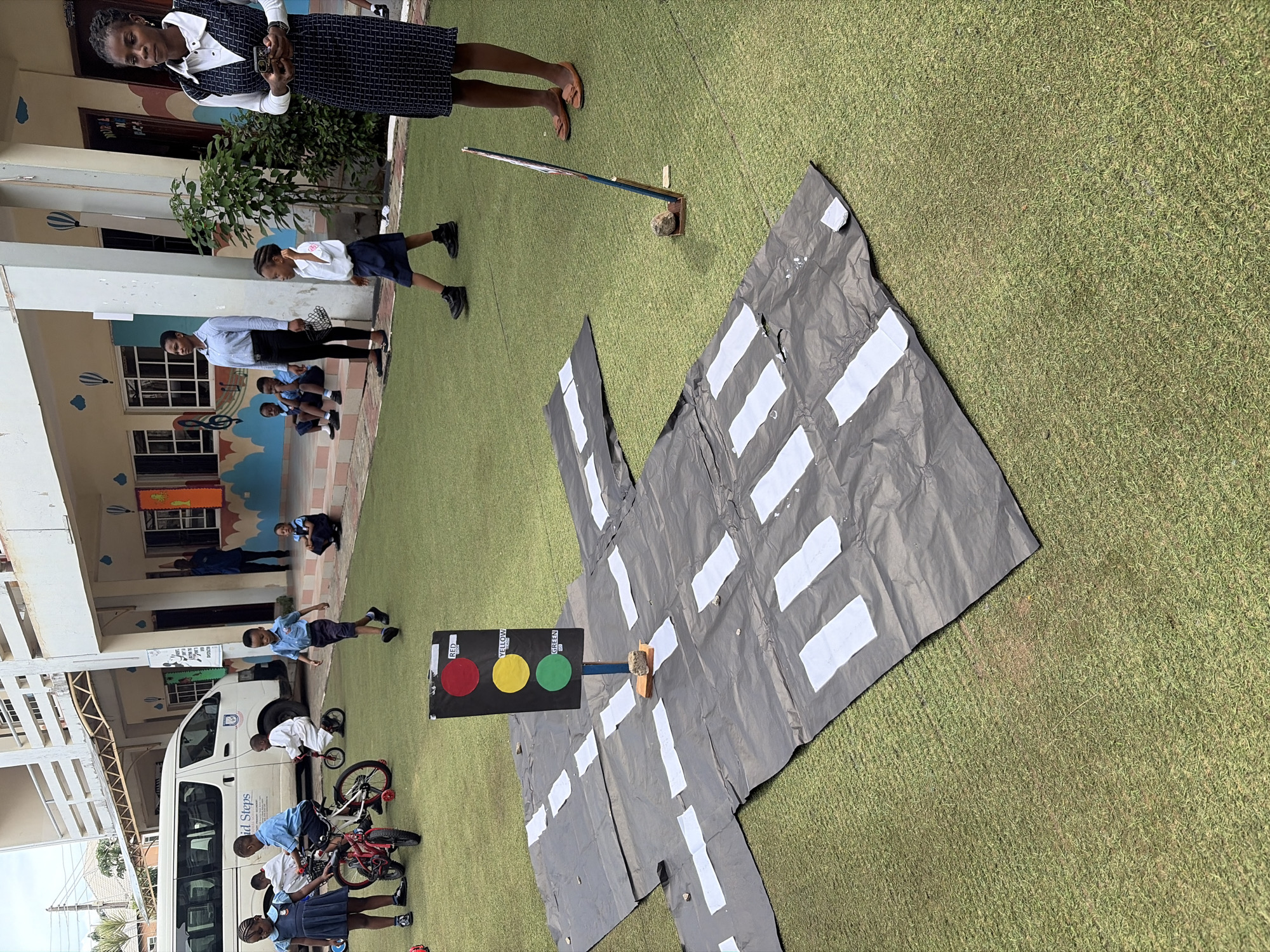 The image shows an outdoor scene in Unknown, Unknown, potentially at a school or community event. A group of children in school uniforms are visible, some with bicycles, and an adult female teacher or organizer is observing. There's a white van parked nearby. A large sheet of dark gray material with white stripes represents a road or intersection, with a homemade traffic light positioned on it. A woman is standing at the top of the frame. The environment is on a grass surface.