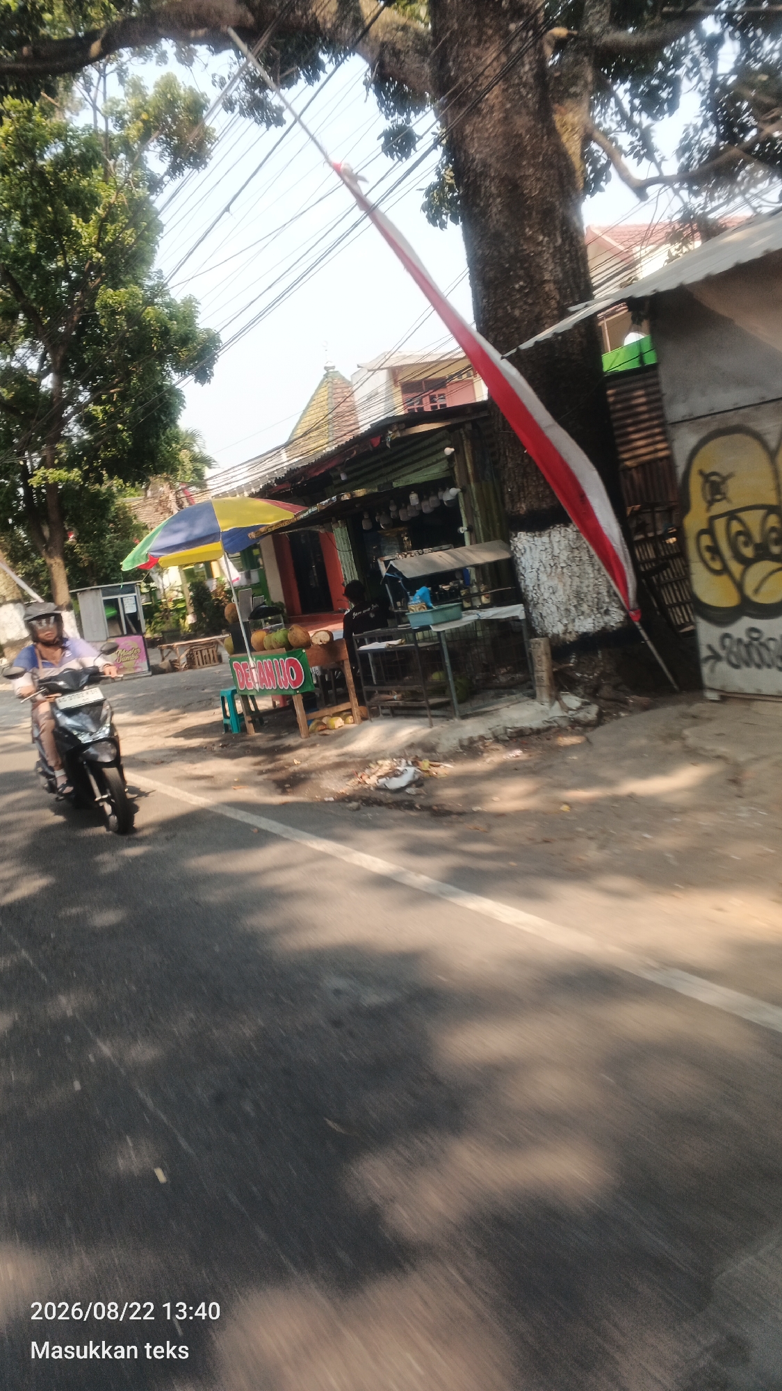 The image captures a street scene in Malang, Indonesia, featuring a person riding a motorcycle on the right side of the road. The rider is wearing a helmet and a light-colored shirt. A roadside stall with a colorful umbrella is visible, selling coconuts. In the background, there are buildings, trees, and overhead power lines. A red and white flag is prominently displayed, attached to a tree or pole, and unfurled at an angle. The road surface shows a white line marking the lane. Graffiti is present on a wall to the right, depicting a cartoon-like character. The overall environment suggests a warm, sunny day, with shadows cast on the road from the trees. The timestamp in the lower left corner indicates "2026/08/22 13:40".