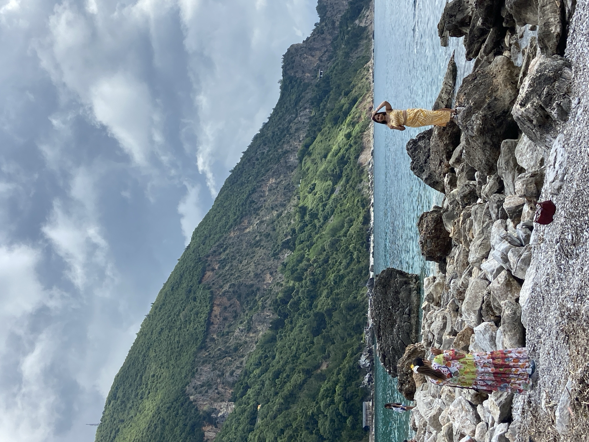 The scene is a coastal view in Prijevor, Montenegro, featuring a rocky shoreline. Two women are present. One is standing on a rock in a yellow patterned outfit, with her hands raised behind her head. The other is on the rocks closer to the foreground, wearing a floral dress and holding a stick. A mountain covered in green vegetation rises steeply from the coastline. The sea is turquoise. The sky is overcast. A rocky shoreline slopes downward with people visible in the lower left. A reddish-brown bag is on the rocks on the right side of the image.