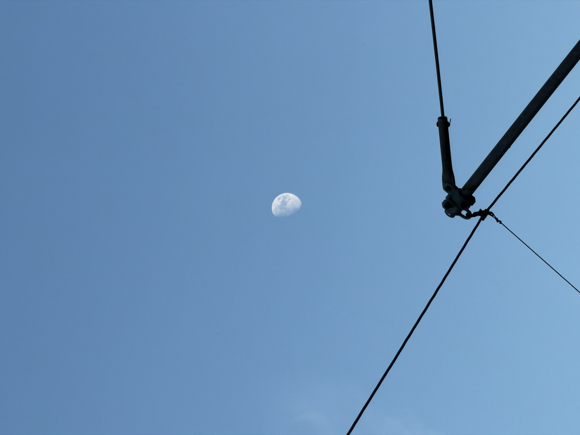 The image is a wide shot of a clear blue sky with the moon visible in the daytime. The moon is a crescent shape and appears to be illuminated from the right. In the foreground, dark electrical wires and poles are silhouetted against the sky, suggesting an urban or transportation setting. The wires create a series of diagonal lines that bisect the frame. The overall impression is one of a serene, clear day, with the moon a celestial object contrasting with the man-made infrastructure. There is no text visible in the image.