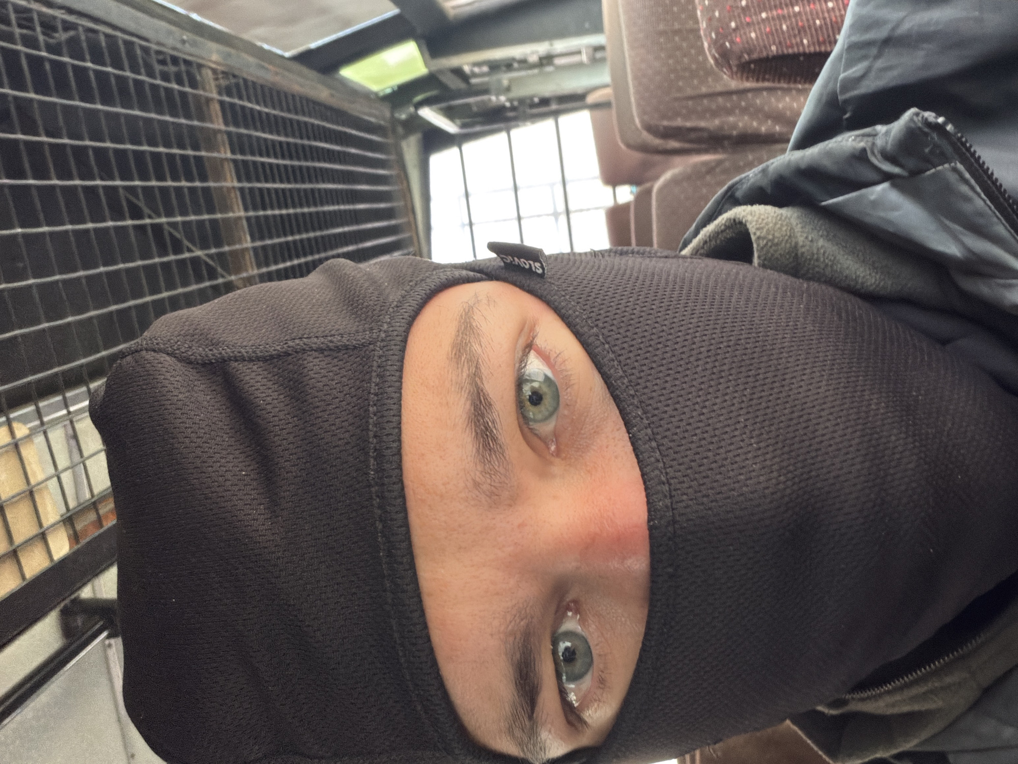 A person wearing a black balaclava and jacket sits on a bus. The balaclava covers their face, exposing only their eyes and eyebrows. The person's eyes are light green. The bus seats are visible in the background, upholstered in brown fabric with a patterned design. The bus ceiling is made of metal bars. The image is a close-up shot of the person's face. The location is not identifiable as Gayk, India.