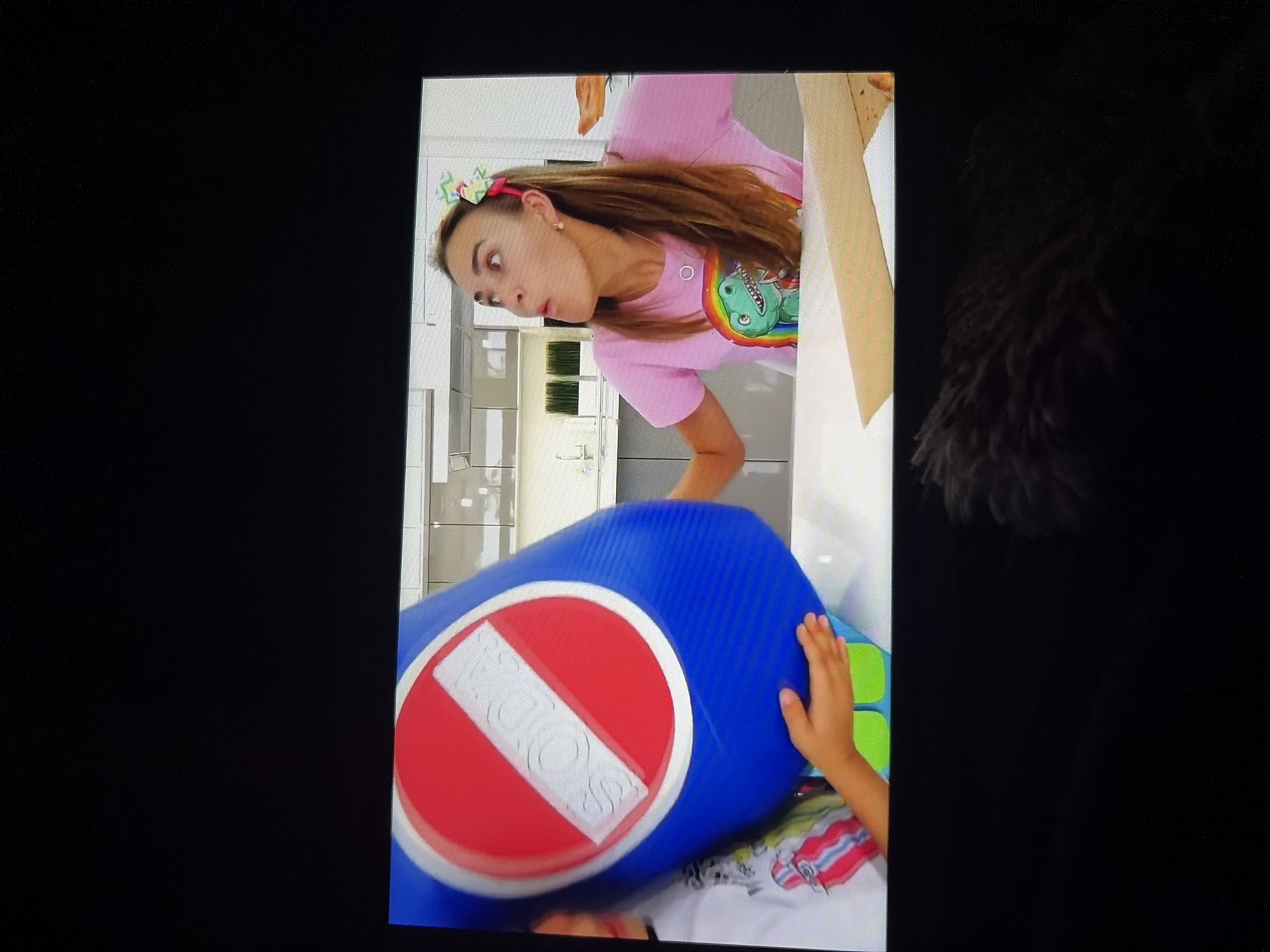 The image shows a woman wearing a pink t-shirt with a dinosaur graphic and a colorful bow in her hair, looking surprised. She is in a room that appears to be a kitchen, with modern cabinets and countertops visible in the background. In the foreground, a large blue object with a red circle and a white diagonal stripe, resembling a "no soda" sign, is being held by a child whose hands are visible. The child is wearing a white t-shirt with colorful stripes. The overall scene appears to be from a video recording, possibly a children's educational program or a vlog. There is no information in the image to suggest any location in Tambov, Russia.