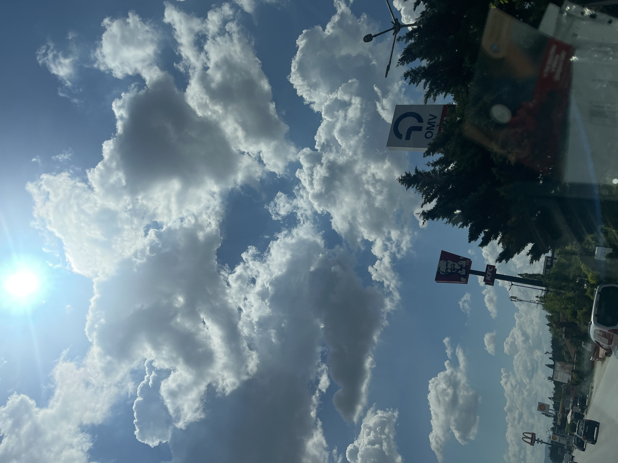 This is a tilted, eye-level shot of a bright, sunny sky with scattered, fluffy white and gray clouds. The sun is visible in the upper left corner, casting a strong lens flare. In the background, on the right side of the frame, there are tall, dark green trees and a street scene with buildings and a road.
Visible signs include a blue and white OMV logo sign, a red and white KFC sign with Colonel Sanders' image, and a McDonald's M logo. The presence of these fast-food chains and the OMV gas station sign suggests a commercial or roadside setting. The time of day appears to be midday due to the bright sun and clear sky. The weather is fair and sunny. The overall mood is bright and cheerful, dominated by the expansive sky.