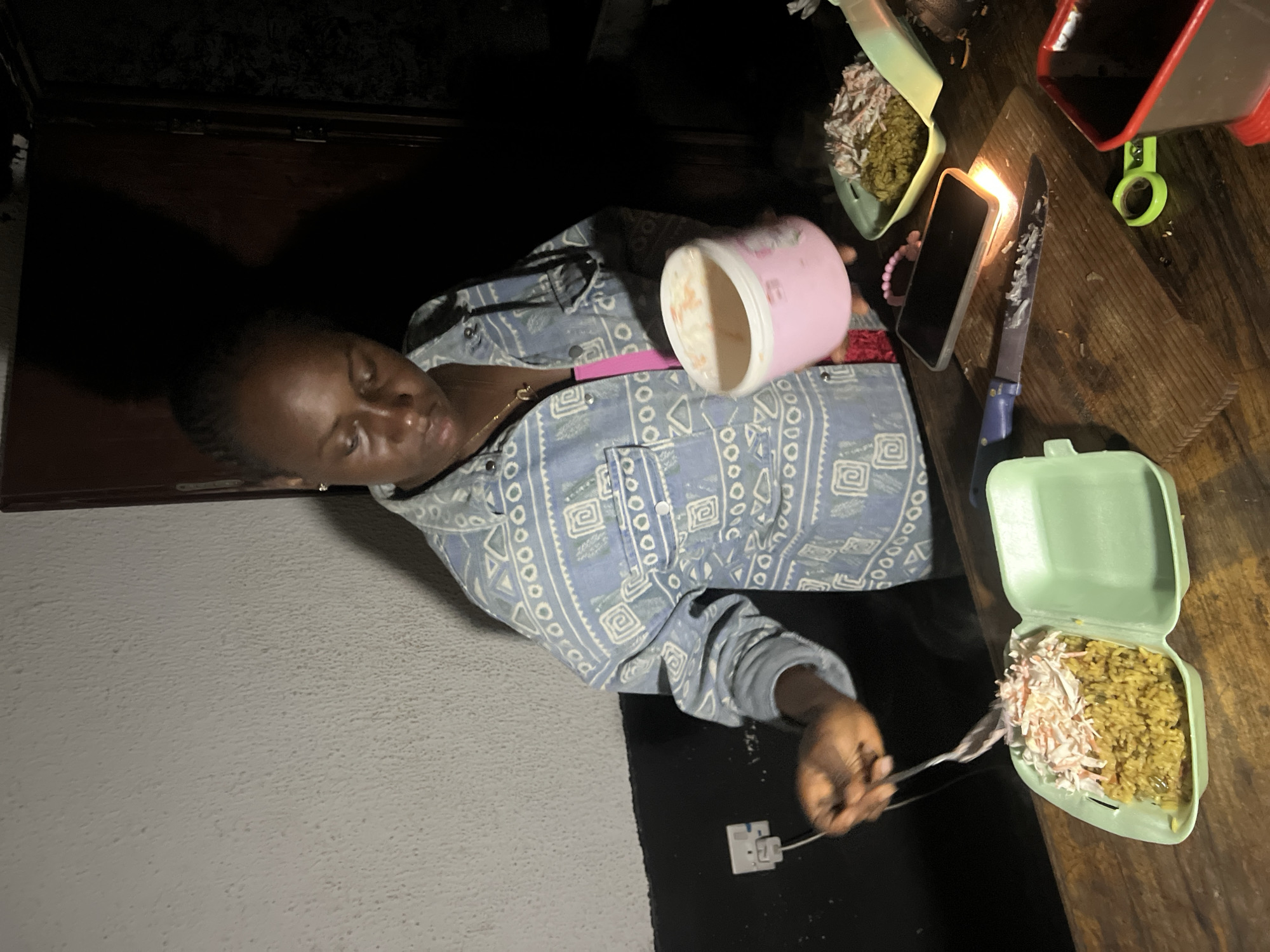 A young woman stands in a dimly lit room, likely in Durumi, Nigeria, preparing food. She is wearing a patterned denim jacket over a pink top. In her right hand, she holds a pink tub of what appears to be a creamy substance, possibly coleslaw or mayonnaise. Her left hand is extended over a green disposable food container filled with rice and topped with shredded ingredients. A fork is held in her left hand, as if she is in the process of serving or mixing the food. On the wooden surface before her, a smartphone is lit by the light of a small, burning object, illuminating a knife with remnants of food on its blade. To the right, another green food container holds a portion of shredded meat or chicken, and a pair of green scissors lies nearby. The background is dark, with a light-colored wall to the left featuring an electrical outlet and a dark door on the right. The overall scene suggests a domestic setting where food is being prepared or served.