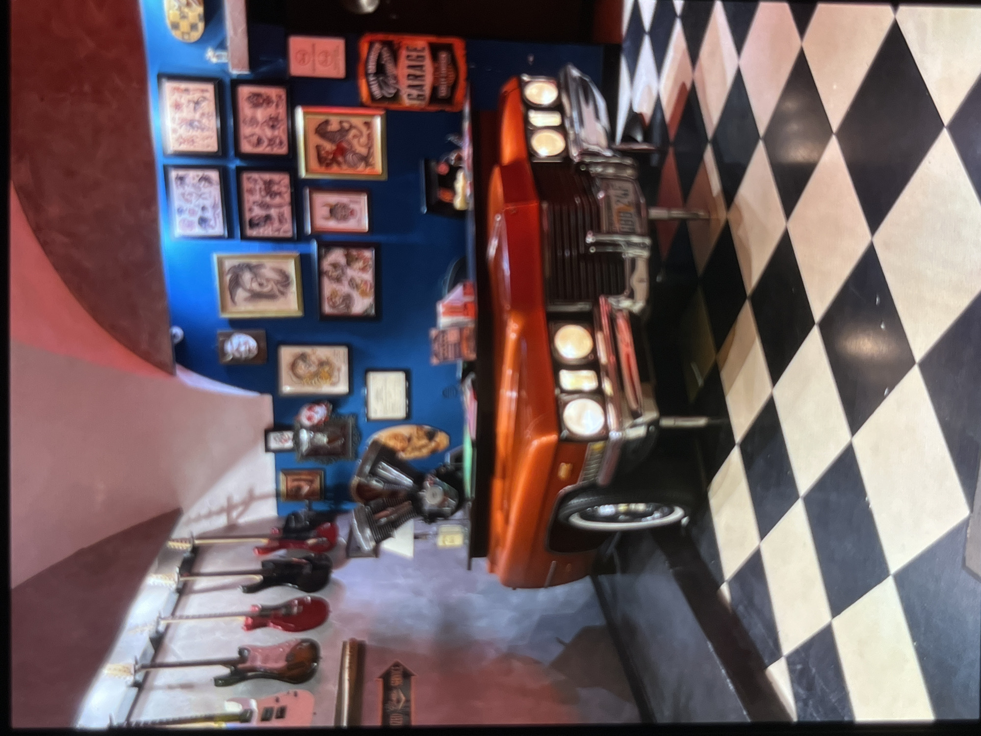 The image shows a room with a checkered black and white floor. In the center of the room is an orange vintage car that has been converted into a reception desk. The car has a chrome bumper and headlights. Behind the car, on a blue wall, are several framed pictures, possibly tattoo designs. To the left of the desk, several electric guitars are hanging on the wall. The room appears to be a tattoo parlor or a similar establishment with a rock and roll theme. There are no people visible in the image. The overall impression is one of a unique and stylish interior design.