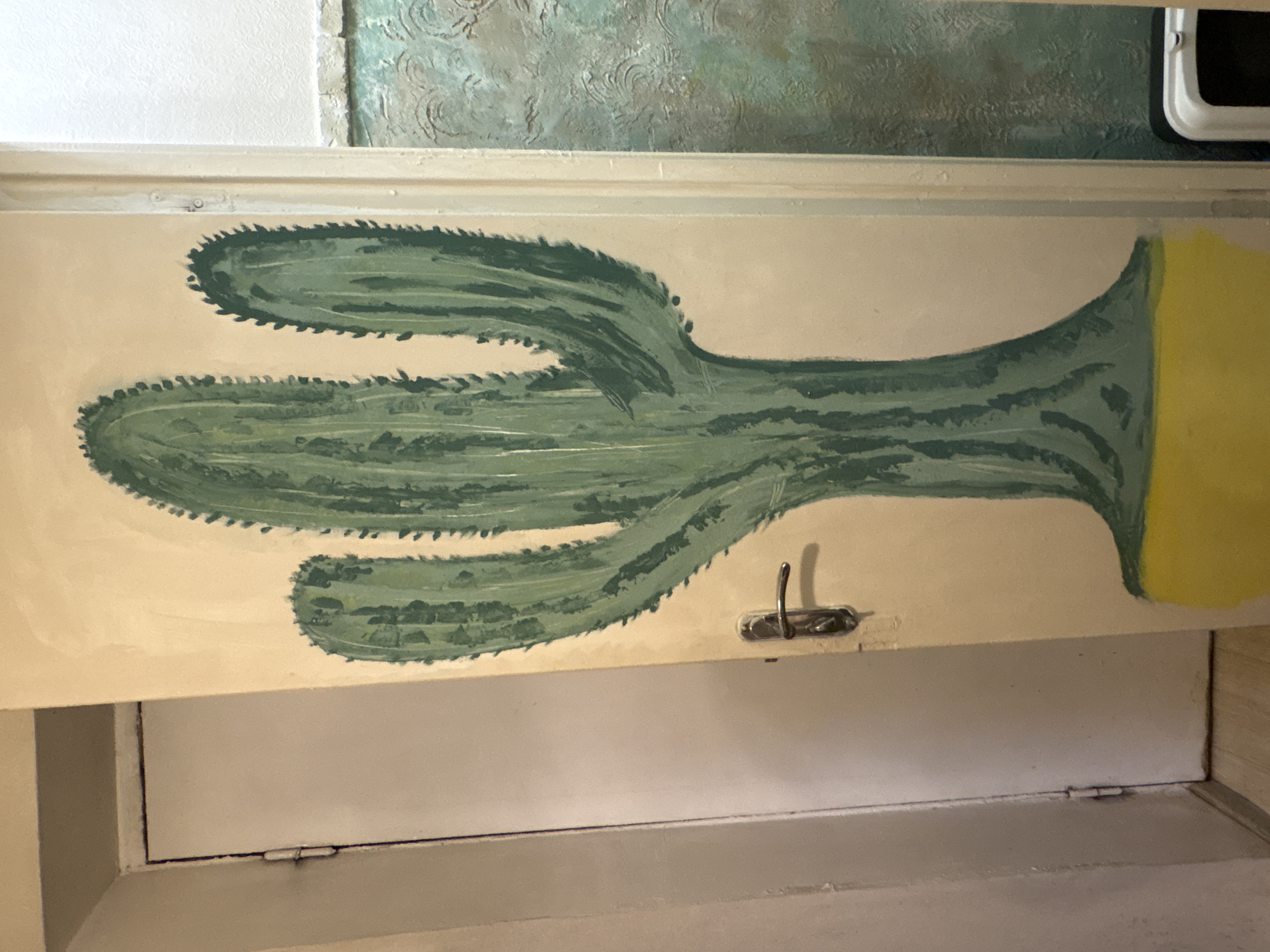 An interior shot depicts a mural of a green cactus on a cream-colored wall. The cactus is tall and has three prominent arms. At the base of the cactus, a yellow ground is painted. To the left of the mural, a white door with silver hinges and a silver door handle is visible. To the right of the mural, a textured green wall is present, and a portion of a white and black object, possibly a bin, can be seen. The image appears to be taken indoors, likely within a residential or public space in Lednevo, Russia.