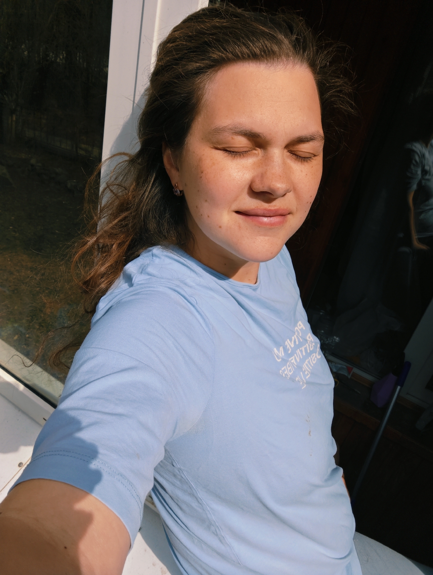 A young woman is standing outdoors in Chaikovsky, Russia. She has fair skin, brown hair, and freckles. Her eyes are closed and she is smiling slightly. She is wearing a light blue t-shirt with white lettering. There is a window behind her to the left. A purple broom and other objects are visible behind her to the right. The lighting suggests it is daytime.