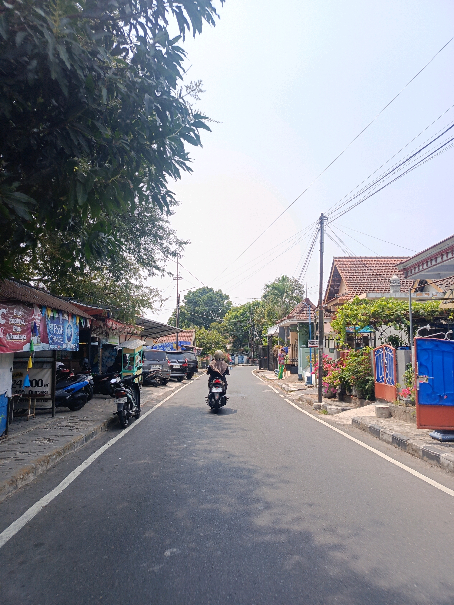 This is a street scene in Tambran, Indonesia. The image shows a road with traffic, including a person riding a motorcycle in the foreground, heading away from the camera. On the left side of the road, there are several small shops with awnings and advertisements. Several motorcycles are parked in front of the shops. On the right side of the road, there are houses with red tile roofs and colorful fences. The sky is clear and bright. The overall impression is a typical street in a residential or commercial area in Indonesia.