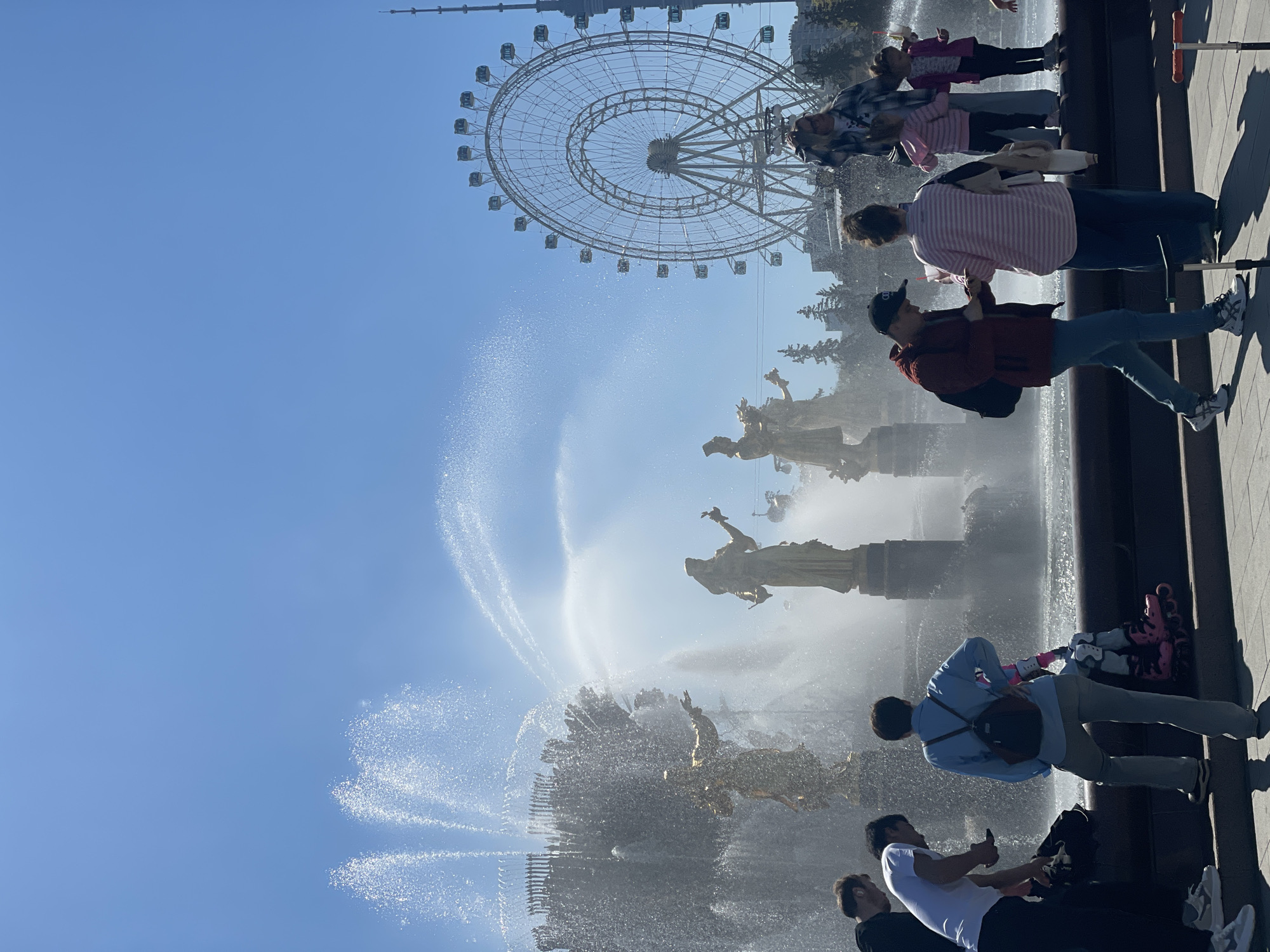 The image captures a scene at the "Friendship of Nations" fountain in VDNKh (Exhibition of Achievements of National Economy), a park in Moscow, Russia. The fountain features a collection of golden statues depicting women representing different Soviet republics. The water jets of the fountain are active and prominent. Several people are visible standing near the fountain, some taking photos. In the background, there is a Ferris wheel. The sky is clear and blue.