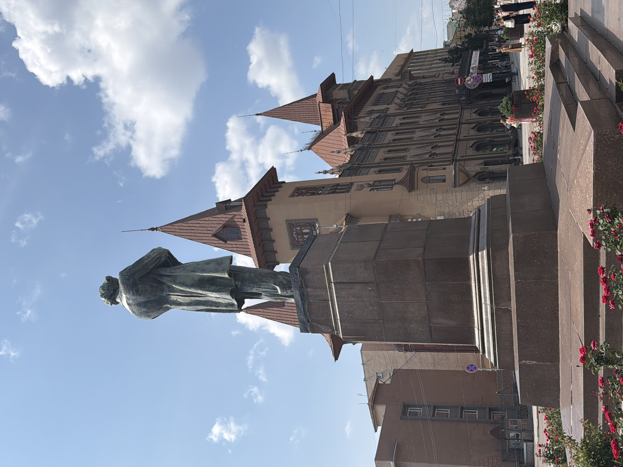 A statue of a man in a coat stands on a stone pedestal in front of a building with a distinctive red-tiled roof. The building appears to be old, with ornate details and multiple pointed towers. Red roses are planted around the base of the pedestal. In the background, there are other buildings, trees, and people walking on a street. The sky is blue with scattered clouds. The image was taken in Saratov, Russia, at the monument to Nikolai Chernyshevsky.