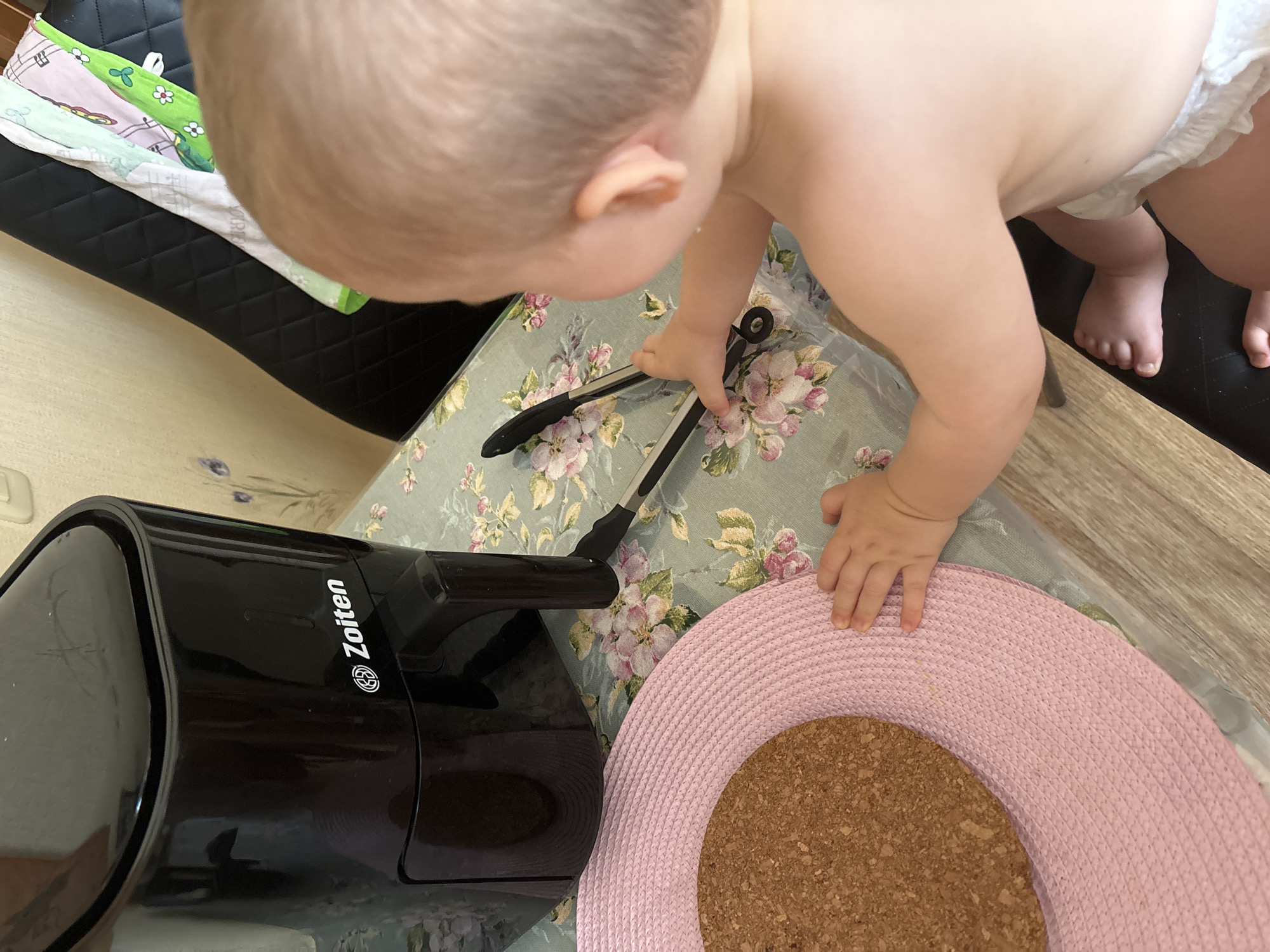 A baby is sitting at a table with a floral tablecloth and a black air fryer. The baby is reaching for a pair of tongs on the table, near the air fryer. A pink placemat with a cork coaster is also visible on the table. The scene appears to be indoors, possibly in a kitchen or dining area. The location of the image is not specified in the provided information.
