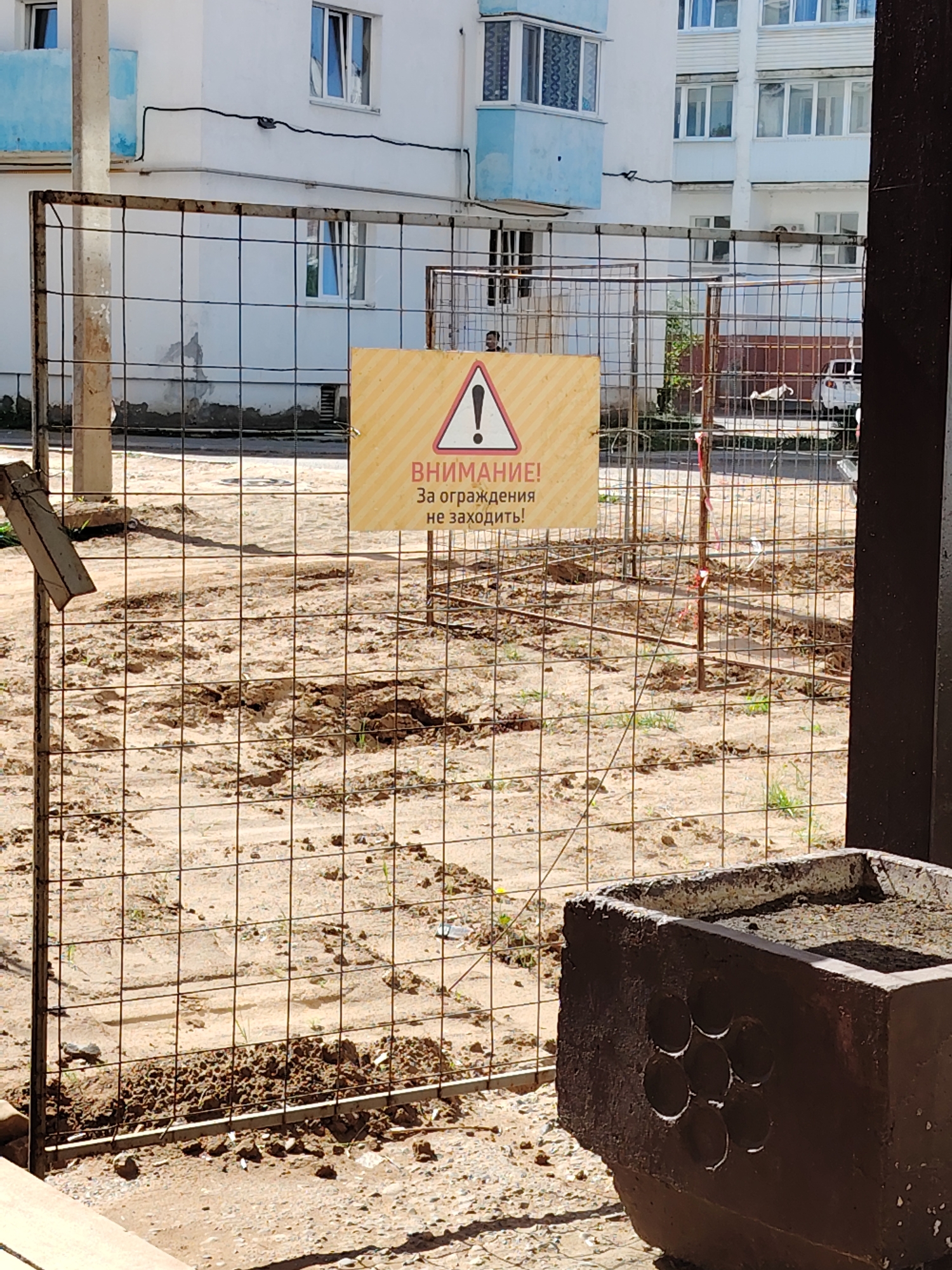 A construction site is fenced off in Chaikovsky, Russia. The ground within the fence is disturbed and muddy. A yellow warning sign with a black exclamation mark and the text "ВНИМАНИЕ! За ограждения не заходить!" ("Attention! Do not enter the fence!") is attached to the fence. A multi-story building stands in the background. A concrete planter is visible in the foreground.