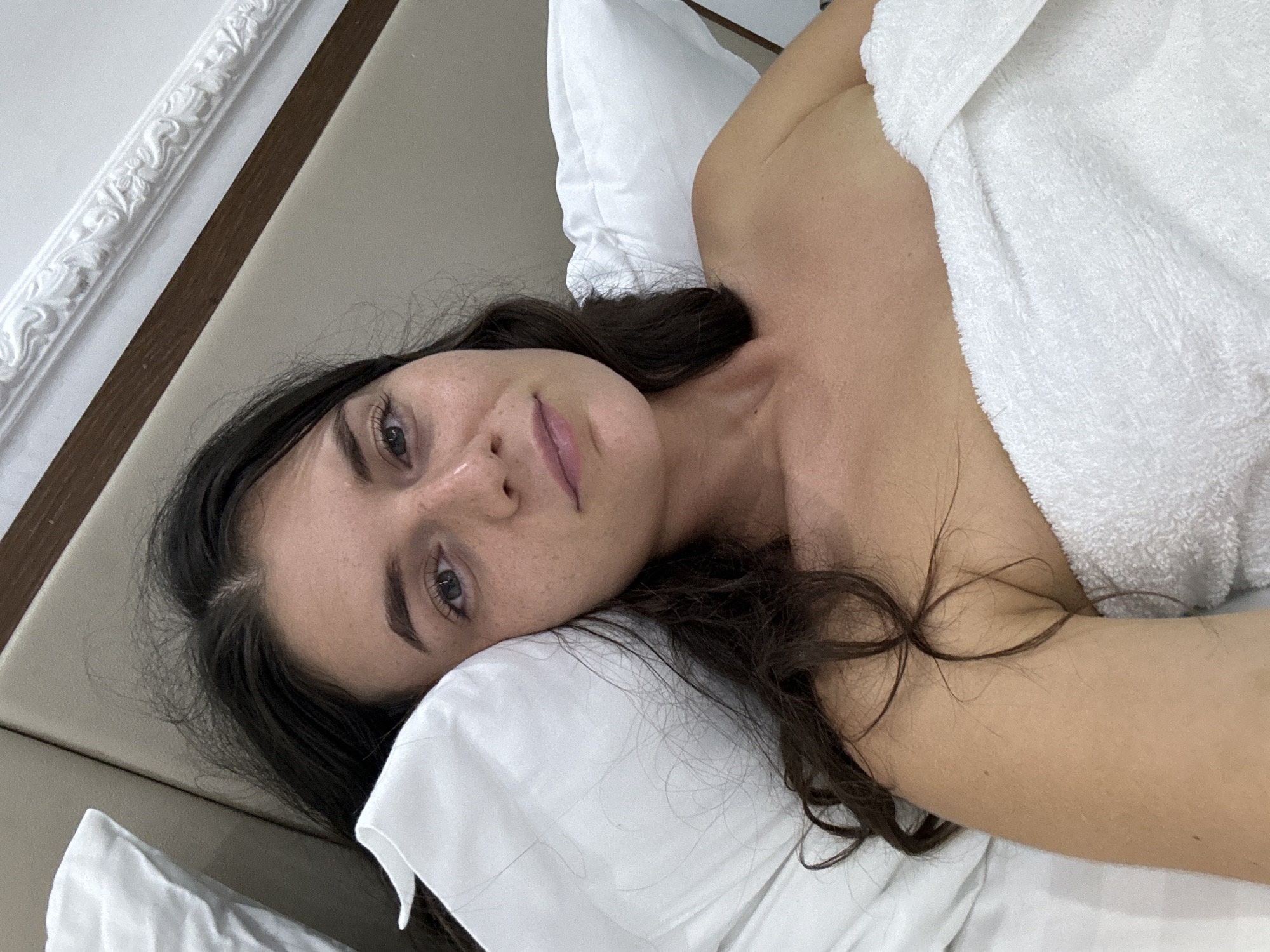 A woman is lying on a bed. She is partially covered by a white towel or blanket. She is resting her head on a white pillow. The setting appears to be a bedroom with a headboard. The location in the city of Thủ Đức, Vietnam, is not determinable from the image.