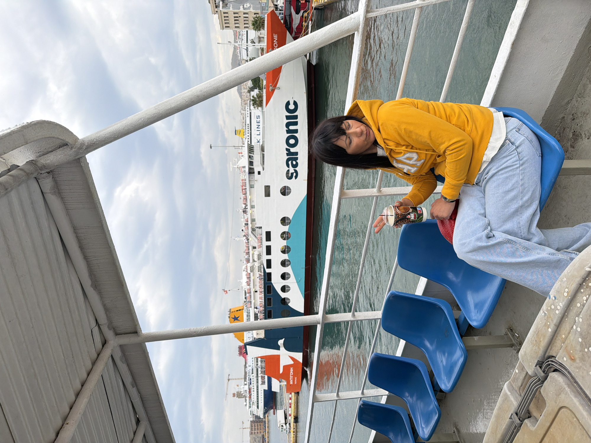 A woman in a yellow hoodie and blue jeans is seated on a blue plastic chair on the deck of a ferry. She is holding a cup with a straw, tilting it slightly. Behind her is the rail of the ferry and the water. Further back, the "Saronic Lines" ferry is docked, with buildings visible in the background. Cranes and another ferry, labeled "Antitoni", are also visible. The location is Piraeus, Greece, evidenced by the docked ferries and port infrastructure.