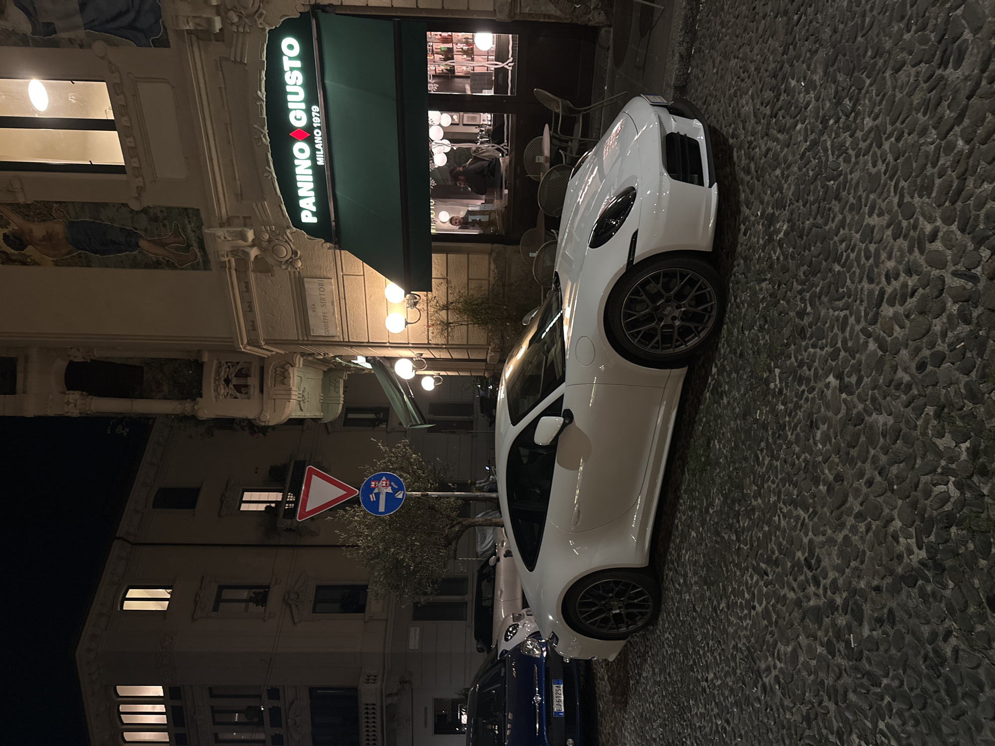 The image is taken at night in Milan, Italy, outside the "Panino Giusto" restaurant. The establishment’s signage is visible, indicating its name and “Milano 1979.” A white car, likely a Porsche, is parked along the cobblestone street. Other vehicles, including a blue van, are also parked nearby. The surrounding buildings are multi-storied and feature architectural details. Streetlights illuminate the scene. People can be seen inside the restaurant through the window. A yield sign and a street sign pole are visible near the parked cars.