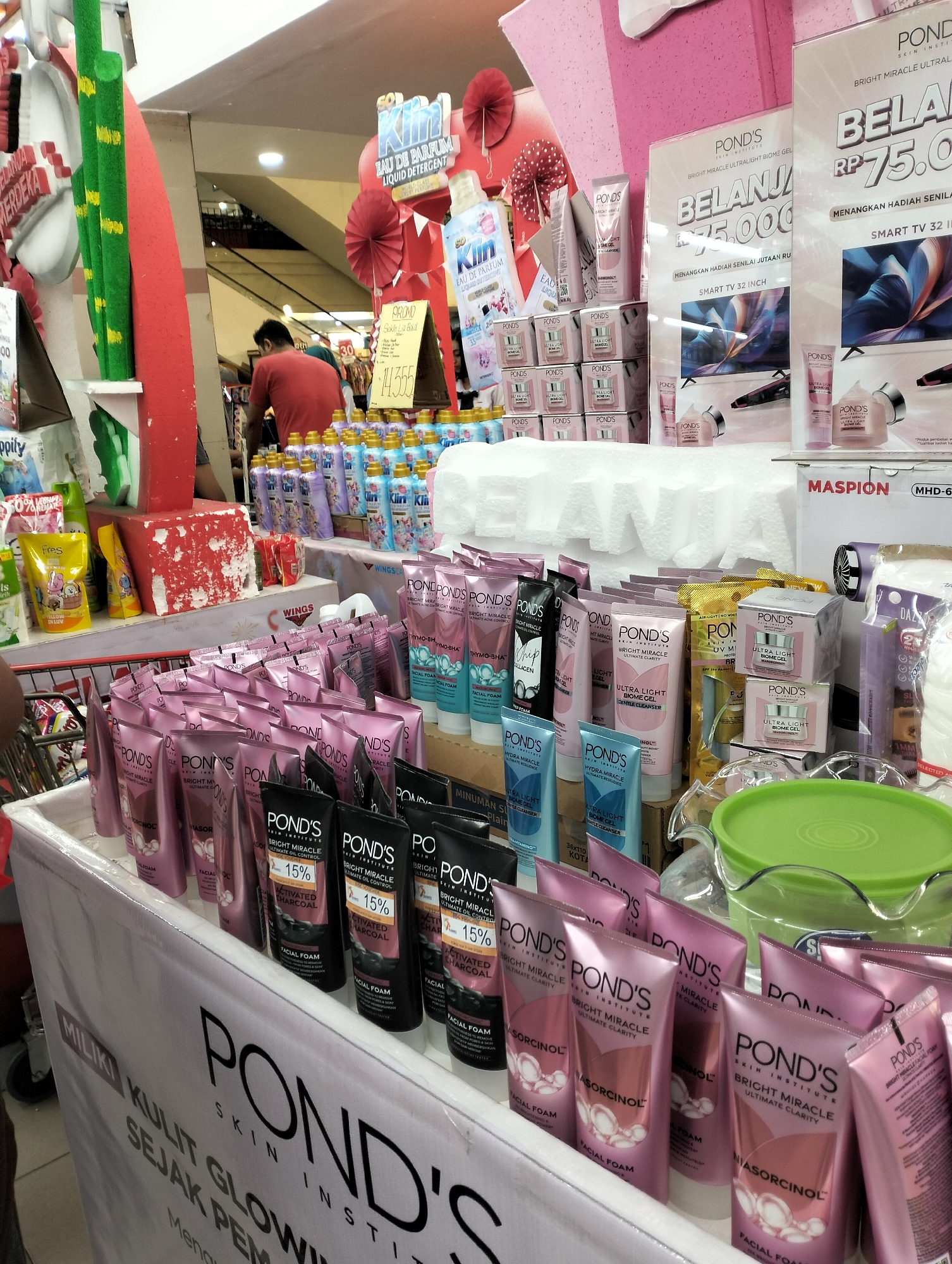 The image depicts a retail display of skincare products, primarily Pond's brand, in what appears to be a supermarket or department store in Ungaran, Indonesia. Several rows of Pond's facial foam and gel products are neatly arranged on tables and shelves. Some products are in pink and black packaging, with labels indicating "Bright Miracle" and "Ultimate Clarity." A large banner in the foreground displays the Pond's logo and promotional text in Indonesian, including "MILIKI KULIT GLOWING" (Have Glowing Skin). Behind the main display, there are other beauty and household items, including bottles of liquid detergent and various smaller packaged goods. A few people are visible in the background, including a man wearing a red shirt who appears to be a staff member or shopper. The overall environment suggests a busy commercial setting with a focus on personal care products.