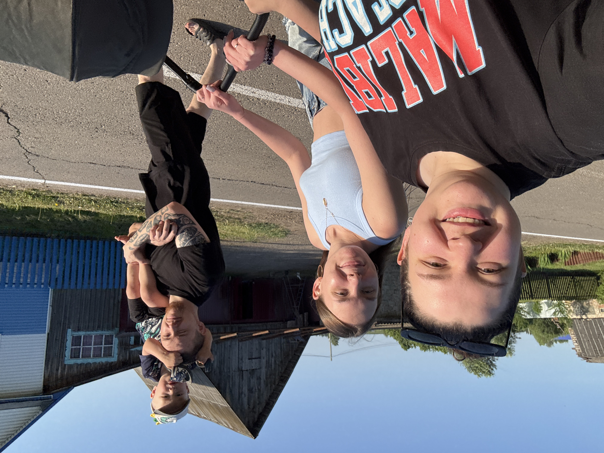 On a sunny day in Lukintsy, Russia, a group poses for a selfie. Three young people are visible, along with a man and a small child sitting on his shoulders. The group appears to be standing on a paved road or path. In the background, there are rural buildings, including houses with painted fences. There is an object resembling a baby stroller partially visible on the upper left side of the image. The group seems to be interacting positively.