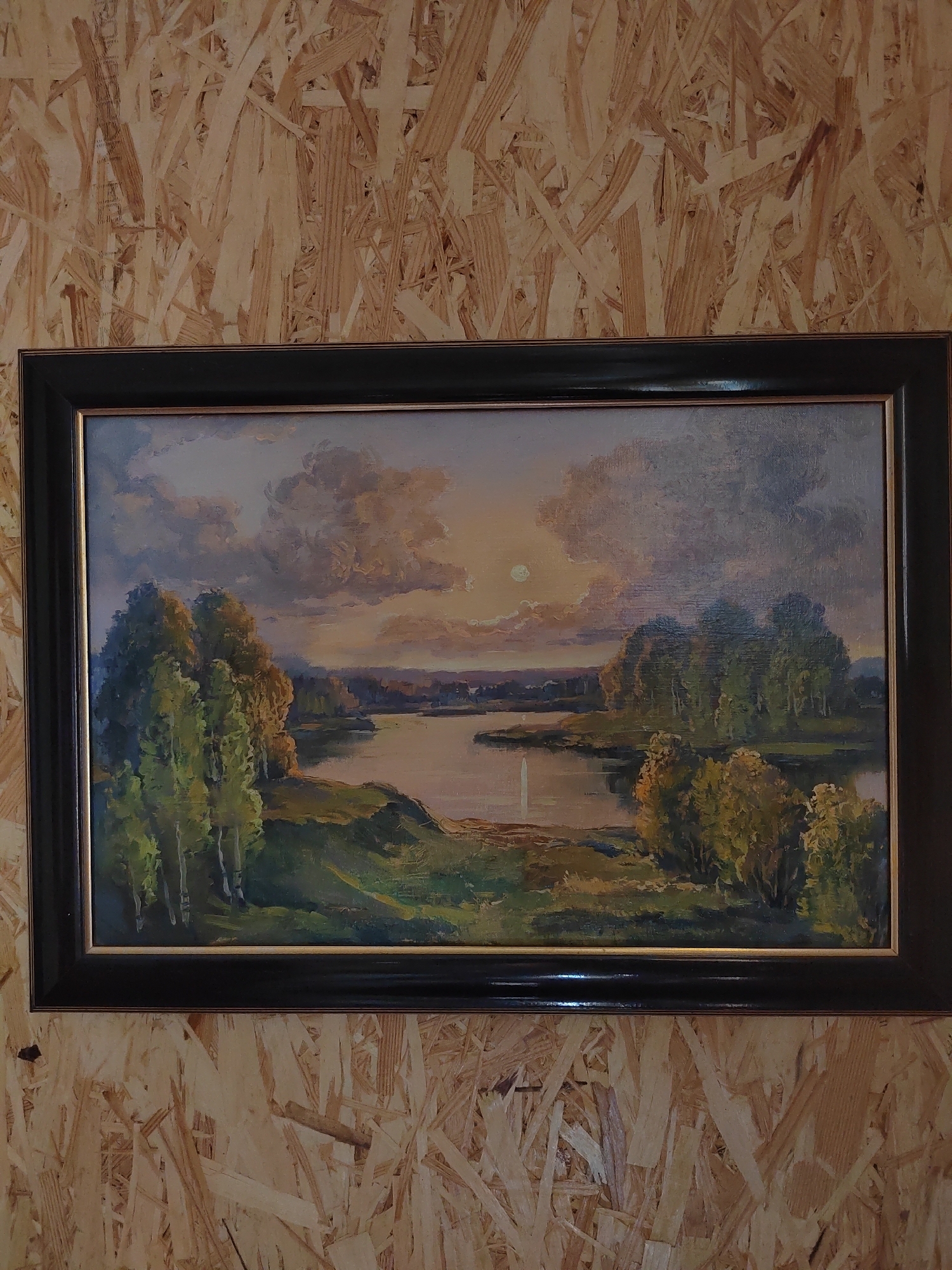 This is a framed oil painting of a landscape, hung on a wall with a wood-chip paneling. The painting depicts a serene scene with a body of water, possibly a river or lake, reflecting the warm hues of a setting sun or moon. Lush green trees and vegetation line the banks, with hints of autumnal colors in the foliage. The sky is filled with dramatic clouds, illuminated by the soft light of the celestial body, which is partially obscured by clouds. The painting is encased in a dark, possibly black, frame with a thin gold inner border. The overall impression is one of tranquility and natural beauty. The location is identified as Бярэзінскі сельскі Савет, Belarus.