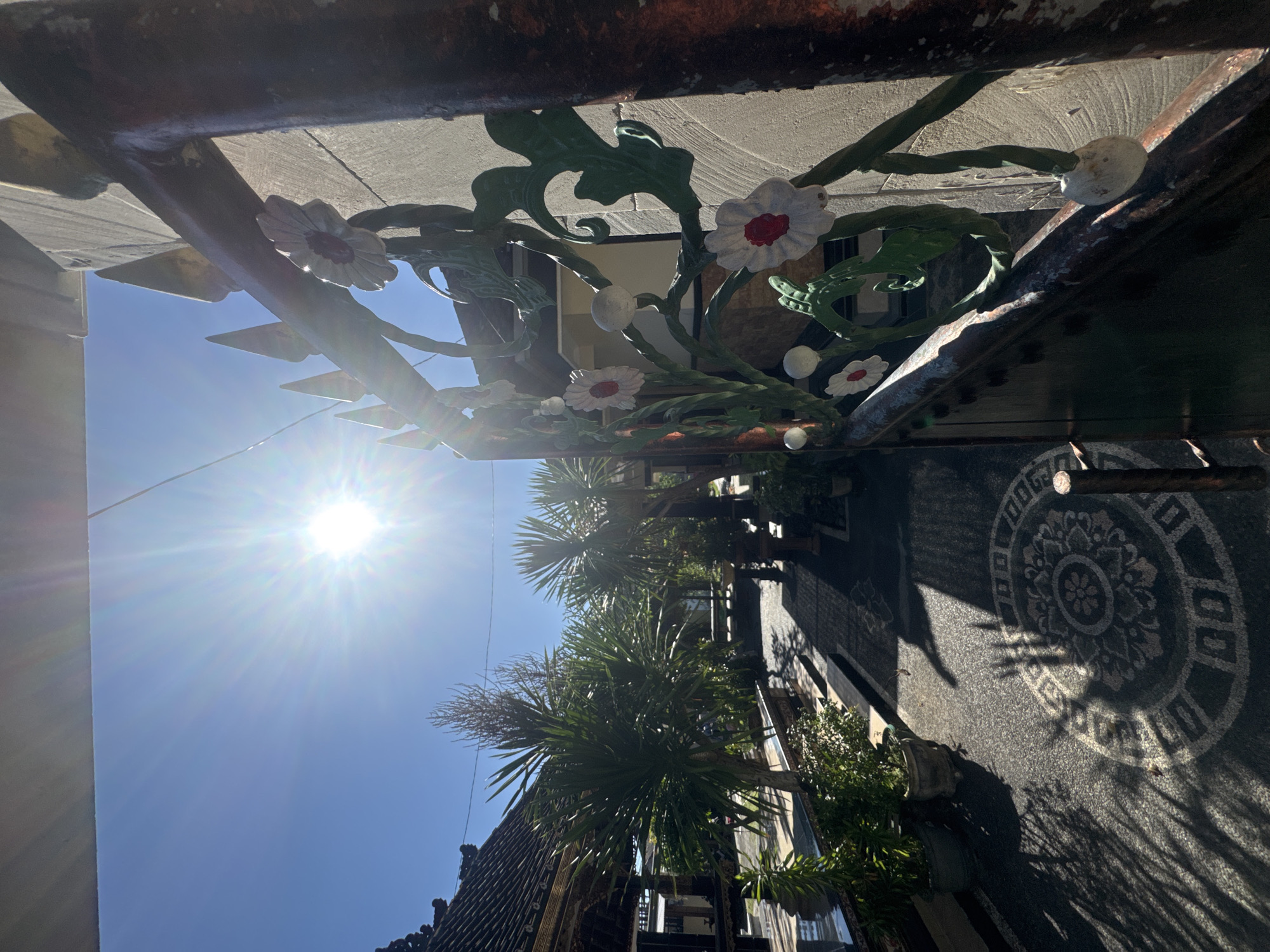 The image shows a street scene in Kuta, Indonesia, viewed from an unconventional perspective. The viewer appears to be positioned under an overhanging structure decorated with ornate metalwork flowers and foliage. A bright sun shines in the blue sky, casting long shadows on the patterned pavement below. Palm trees line the street, adding a tropical feel to the scene. Buildings with tiled roofs are visible along the side of the street.