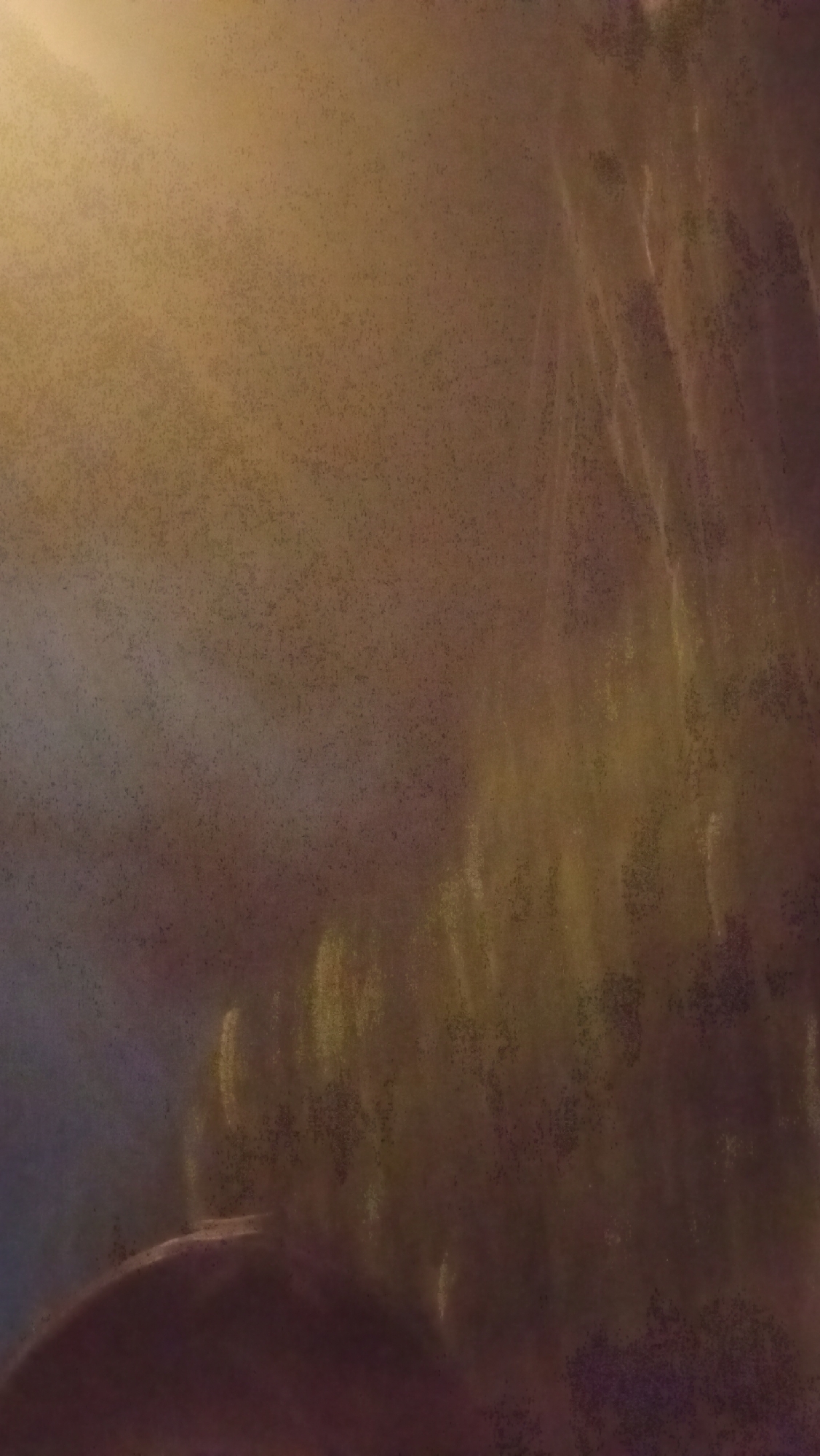 The image is a blurry, abstract shot taken at night, possibly in Kuta, Indonesia. The foreground is dominated by a dark, curved shape that could be a rock or a mound of earth. Behind this, the background is a wash of muted colors, primarily shades of brown, yellow, and purple, with streaks of light that suggest foliage or trees swaying in the wind, possibly illuminated by streetlights or other artificial sources. The overall impression is one of movement and a lack of clear focus, creating an atmospheric rather than a detailed depiction of a specific location. There are no discernible people or specific objects that can be identified with certainty.