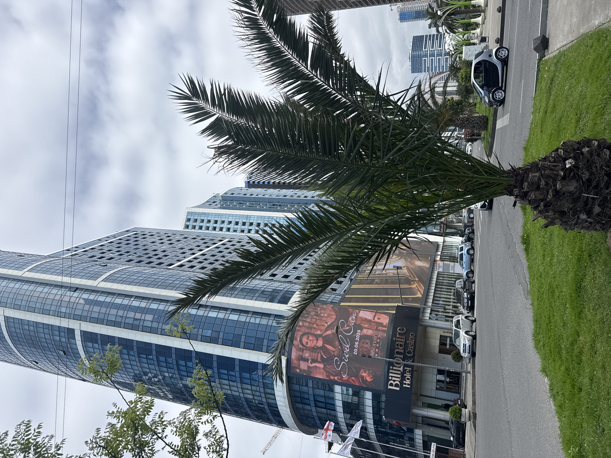 An outdoor, eye-level shot captures a modern urban scene dominated by a tall, glass-clad building under an overcast sky. A large palm tree with lush green fronds occupies the right foreground and extends towards the center, its rough trunk visible at the bottom right. To the left of the palm tree, a wide street with light grey asphalt runs parallel to a sidewalk and a strip of green grass. A small, grey-and-white two-seater car (resembling a Smart car) is parked on the street on the right side of the image, while other cars are partially visible further down the road and parked near the building.
The prominent building, with its blue-tinted reflective windows and angular architecture, curves slightly and has visible horizontal white bands. A large billboard is affixed to the lower section of this building, featuring a woman (likely a singer) and text that reads "Sibel Can," "20.06.2026," "Billionaire Hotel & Casino," "Live in Concert," and "The Grand Casino." Below the billboard, the building's entrance also displays the name "Billionaire Hotel & Casino." Flags, including one identifiable as the flag of Georgia (the country), are visible near the building's base. The sky is largely white and cloudy, suggesting an overcast day. In the distance, other high-rise buildings are visible. The overall scene depicts a contemporary city street, likely in a region with palm trees, such as Georgia.