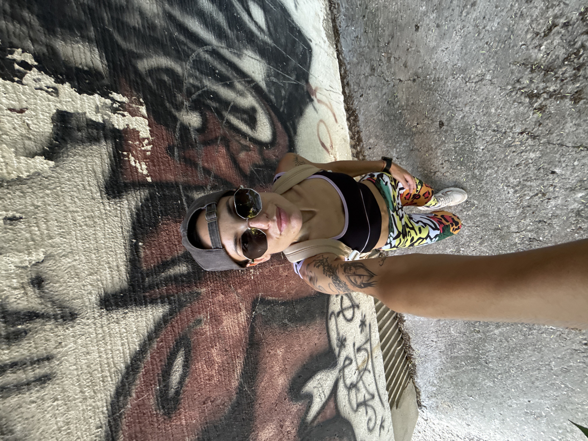 A woman with sunglasses and a grey cap is photographed in an urban setting. She wears a black top with purple trim and patterned leggings. Her arm is extended, presumably holding the camera. Graffiti adorns a wall to her left, while a concrete surface is visible on her right. The setting is possibly Pleven, Bulgaria, but there is no explicit information to confirm this.