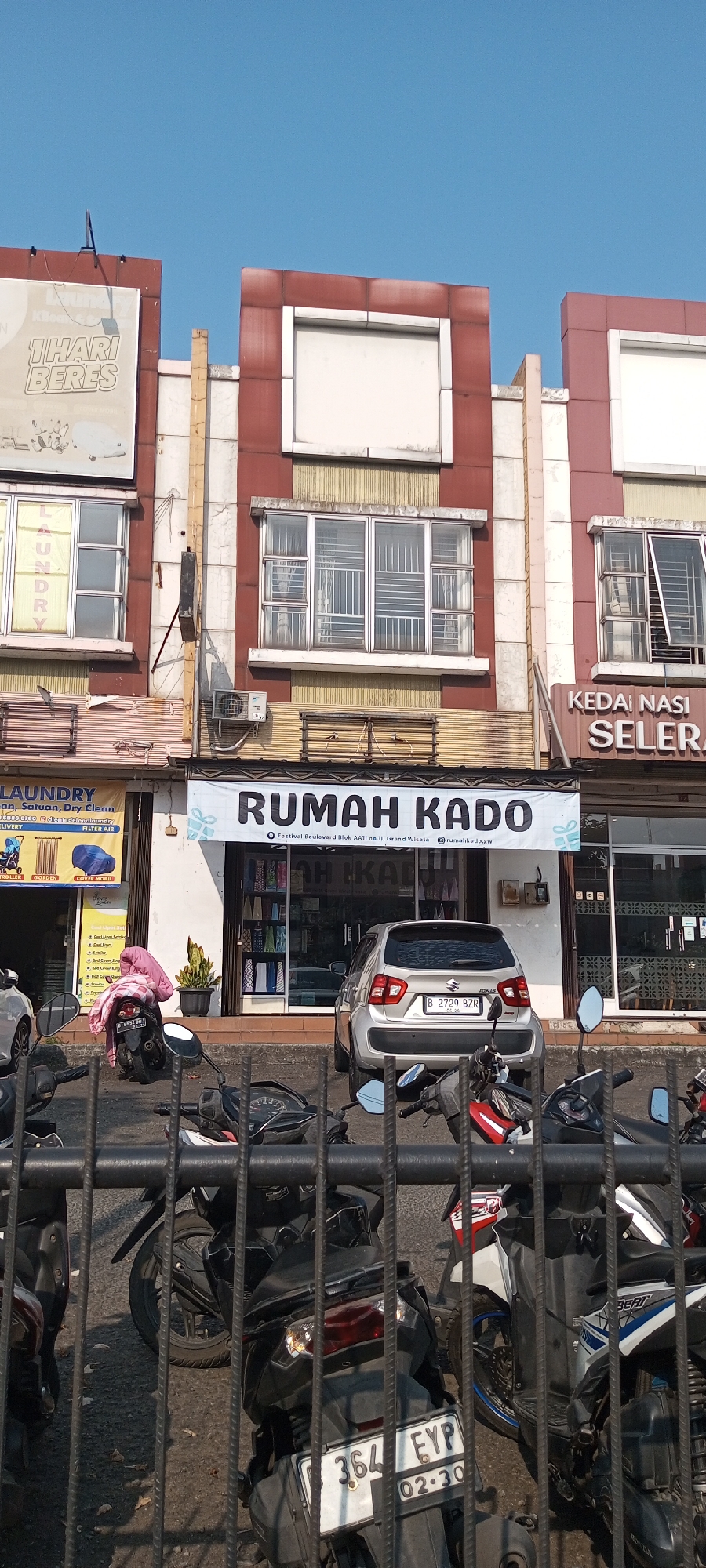 The image shows a street view in Lambangjaya, Indonesia, on a clear day. In the foreground, a black metal fence partially obstructs the view of several parked motorcycles. Behind the fence, a silver Suzuki Ignis car is parked in front of a building. The building has multiple storefronts. One storefront has a banner that reads "RUMAH KADO" in large black letters. To the left of "RUMAH KADO," a laundry service storefront is visible, with a sign that says "LAUNDRY" and lists services. The building's facade is a mix of red and white sections, with large windows and some architectural detailing. A small potted plant is situated near the entrance of the "RUMAH KADO" store. The sky above is a clear blue.