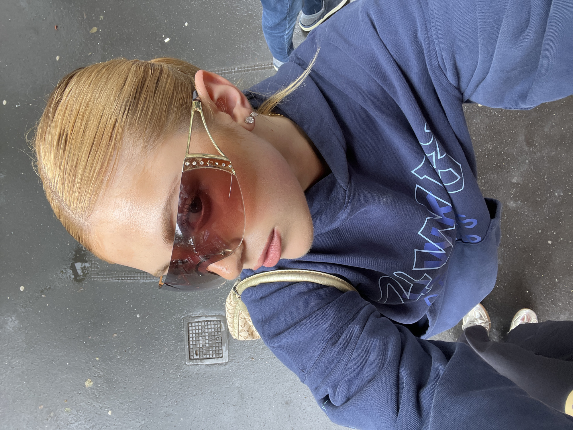 A blonde woman wearing rose-colored sunglasses and a blue hooded sweatshirt is standing on a wet street in Paris. She is holding a beige purse and wearing shiny silver sneakers. The street is gray, and there is a metal utility cover in the foreground. Another person is standing behind her wearing jeans and blue shoes.