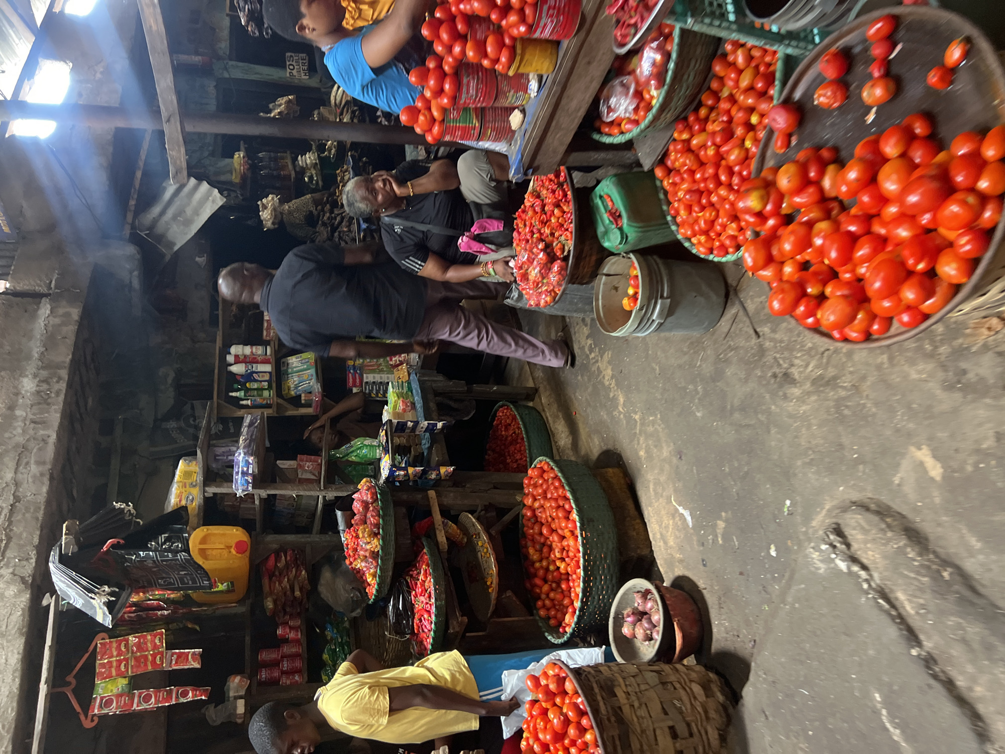 The image shows a bustling scene at a market in Nkpor, Nigeria. Baskets and trays overflowing with tomatoes dominate the foreground and right side of the image. At the top left, a person in a blue shirt sits near stacked containers filled with tomatoes. An older woman in a black shirt rests her head on her hand nearby. A person in a black shirt and tan pants walks towards a store on the left side of the image, where shelves are stocked with various products. A person in a yellow shirt is positioned at the bottom left, attending to the tomatoes. The environment appears to be a covered market area with dim lighting and concrete flooring. Additional items such as onions in a bowl, miscellaneous packaged goods, and containers are visible in the background, suggesting a typical market environment.