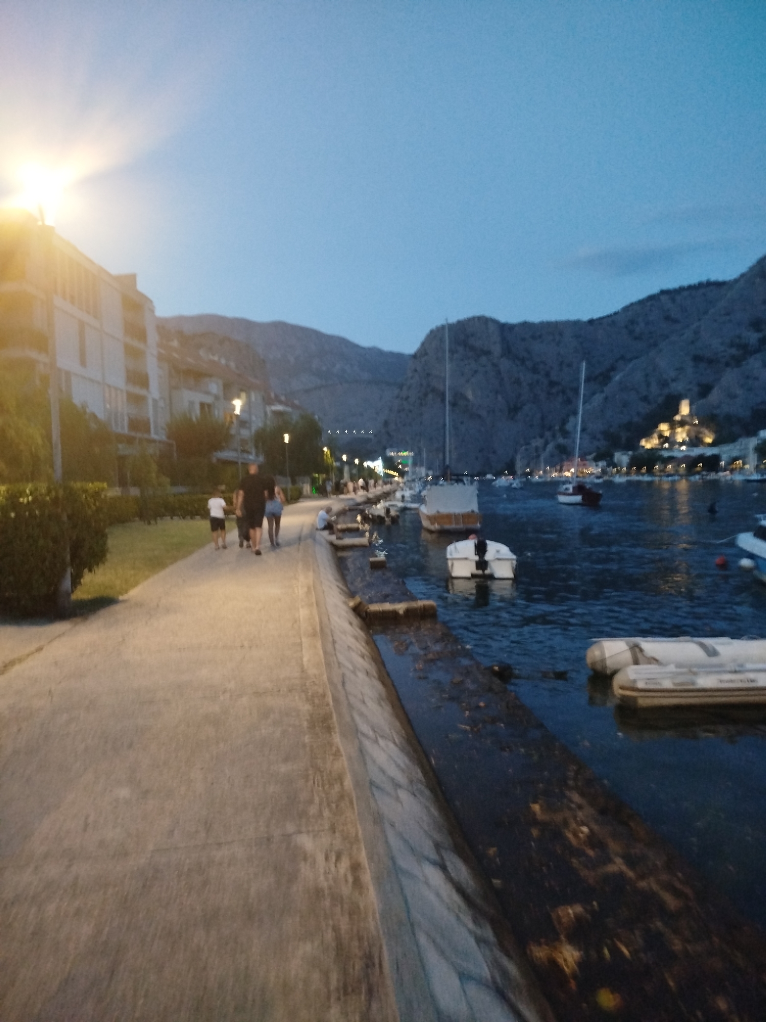 The image depicts a waterfront promenade in Omiš, Croatia, likely during the early evening or dusk. The promenade is paved with concrete and runs alongside a body of water where several boats are moored. On the left side of the promenade are buildings, including what appears to be an apartment complex, and a line of trees or bushes. The sky is a deep blue, suggesting twilight. In the distance, mountains are visible, with a fortified structure, likely a castle or fort, illuminated on one of the hills.
Several individuals are walking along the promenade. Closest to the camera, a group of three people, two adults and a child, are walking away from the viewer. Further down the promenade, other figures can be seen walking.
The water contains various types of boats, including what appear to be dinghies and larger vessels with masts. Some boats are closer to the shore, resting on the edge or in shallow water. The water itself is dark and reflects the surrounding lights. The embankment along the water's edge is made of stone or concrete. The overall scene suggests a leisurely atmosphere in a coastal town.