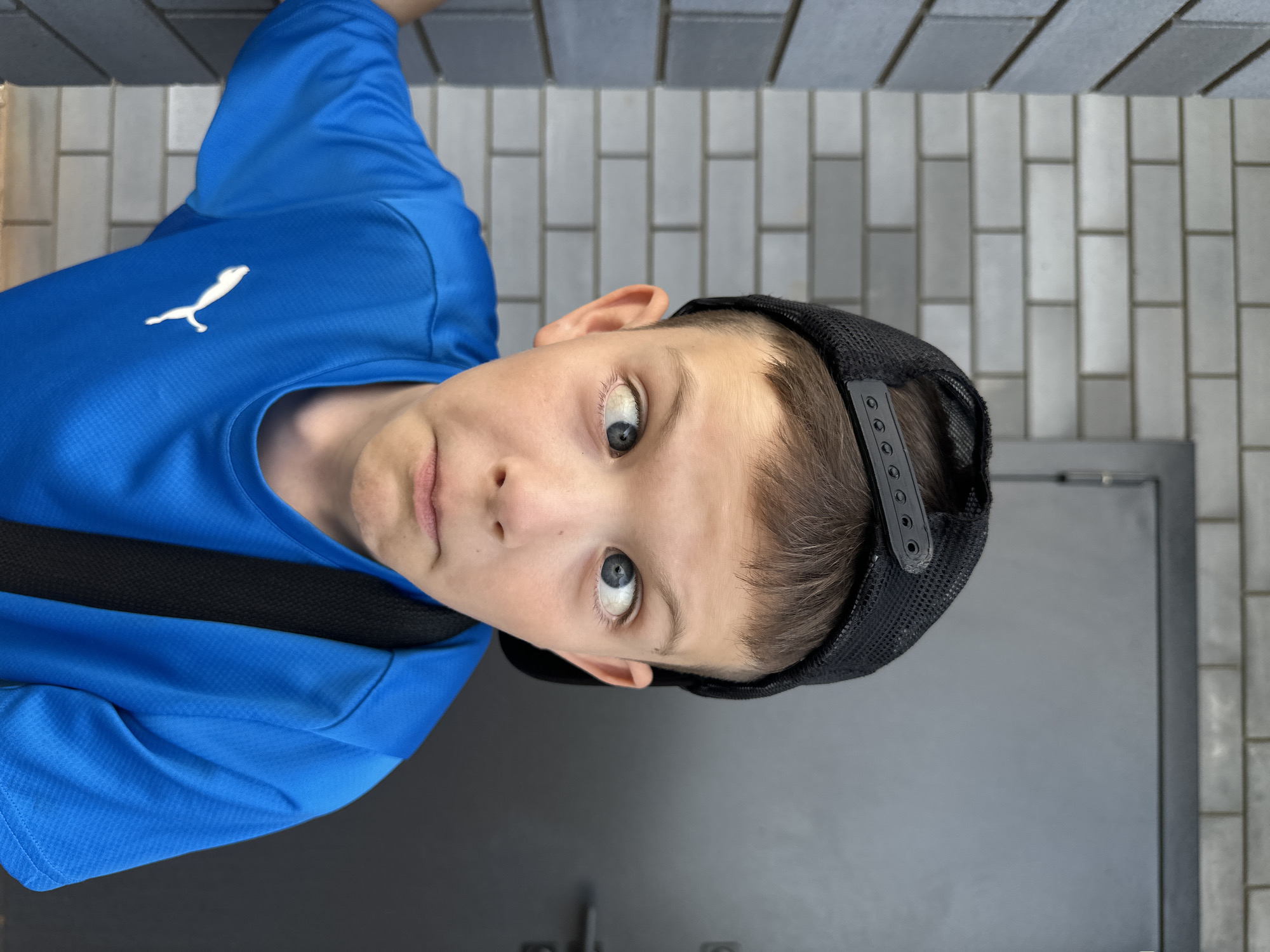 A young boy wearing a blue Puma shirt and a black baseball cap is looking up at the camera. He is standing in front of a light gray brick wall and a dark gray door, potentially in Прикамский, Russia. The boy has light skin and blue eyes. He is wearing a black strap, possibly of a bag, across his chest. The image appears to be taken from a slightly elevated position, looking down at the boy.