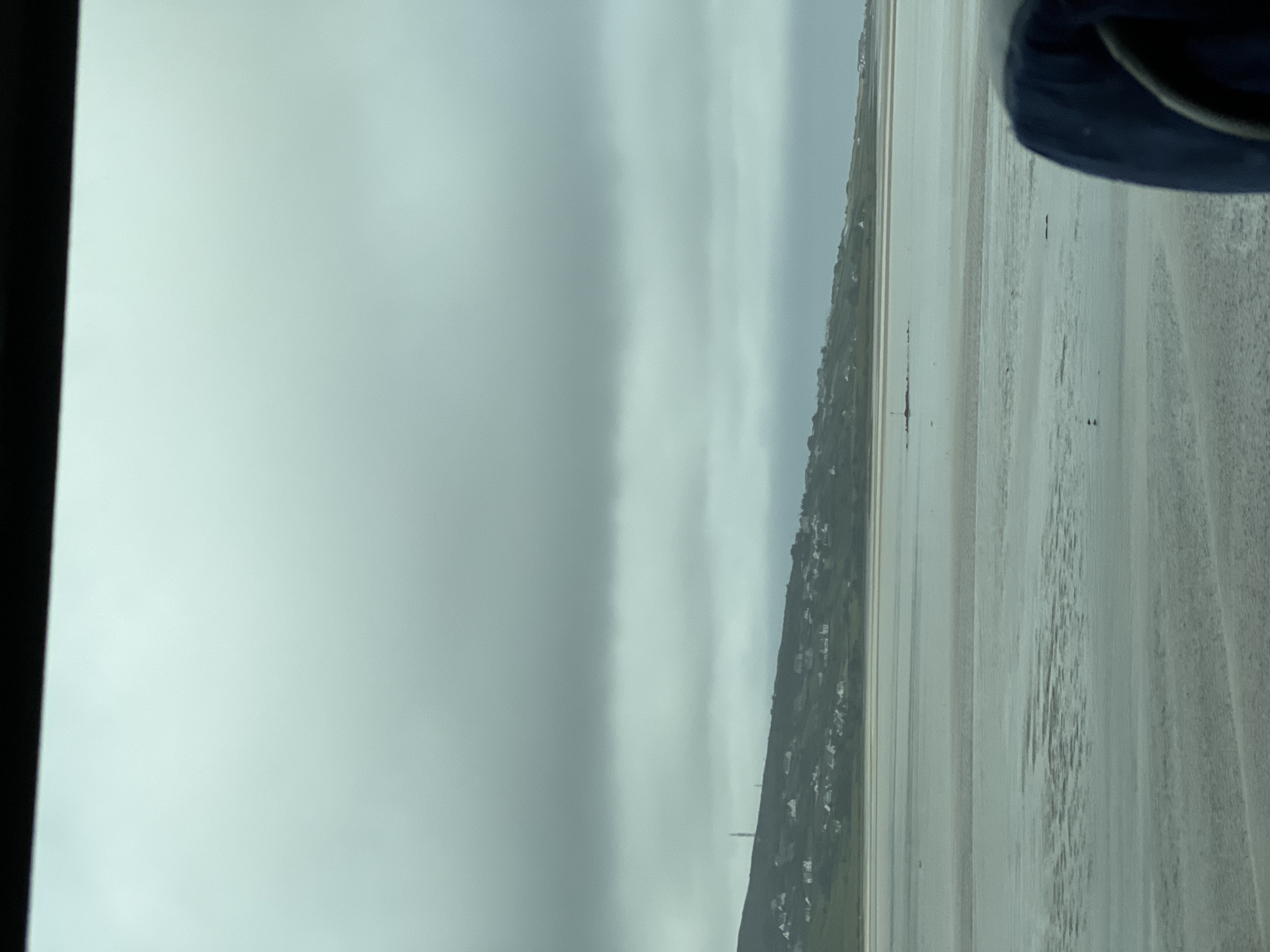 This media file depicts a landscape scene. The dominant subjects are a vast expanse of wet sand, likely a beach at low tide, and a distant, overcast sky. A green, hilly terrain with scattered buildings is visible along the left side of the frame, suggesting a coastal village or town. The sand has parallel, elongated textures, possibly from receding water or wind patterns.
There are no clearly discernible people or specific objects that stand out as the primary focus, though there are some small dark shapes on the sand that could be distant figures or debris. A dark blue object, possibly a piece of clothing or equipment, is visible in the upper right corner.
The weather appears to be overcast and possibly humid, contributing to the muted color palette of the scene. The time of day is indeterminate due to the lack of direct sunlight and shadows. The overall mood is calm and somewhat somber, due to the gray skies.
The location context provided is Killult Middle, Ireland. This information, combined with the visual cues of the coastline and the architecture, helps to place the scene. There is no visible text in the image. The image is oriented vertically, with the top of the frame being the left side in a typical horizontal view.