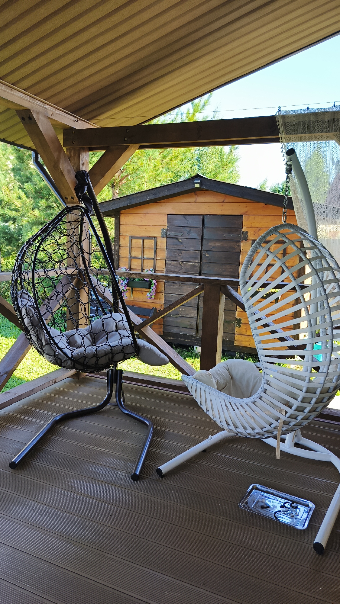 The image shows a covered wooden porch with two hanging chairs. One chair is black and made of woven material with a gray cushion. The other chair is white, also made of woven material, and has a cream-colored cushion. A small wooden shed with double doors is visible in the background, with a window and flower box on its front. The porch floor is made of dark wooden planks. A metal tray with what appears to be some sort of metal hardware is on the porch floor in front of the white chair. The scene suggests an outdoor recreational space, possibly a backyard or garden area. The location is specified as Сокуры, Russia, indicating this is likely a residential property. There are no people visible in the image.