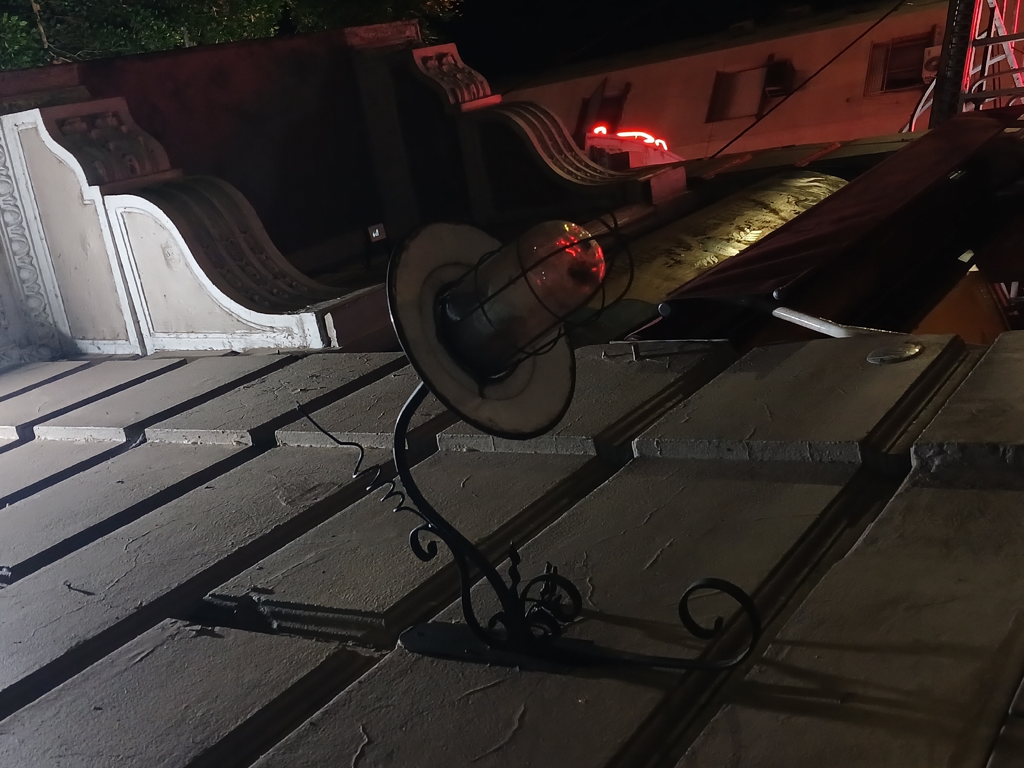 This media file captures a nighttime scene, likely in an urban setting, with a focus on architectural details and artificial lighting. The foreground features a decorative, dark metal wall-mounted lamp with ornate, wrought-iron scrollwork at its base. The lamp has a glass enclosure around its bulb, which appears to be reflecting a reddish light from an external source, giving the impression of an internal glow. The lamp is positioned on a textured, ridged surface, possibly a parapet or roof terrace, made of what looks like concrete or stone slabs.
In the mid-ground, behind the lamp, stands an ornate architectural element, possibly a cornice or facade, characterized by decorative corbels and sculpted details in a light, off-white hue. Beyond this, in the background, a building wall is visible, illuminated by a strong red glow from neon signs. Parts of the neon signs are visible, and the letters "LA" can be clearly discerned in red neon on the building. Other details on the building include windows, an air conditioning unit, and what appears to be a dark, folded awning or canopy. A ladder-like structure is also partially visible on the right.
The overall atmosphere is dark, indicating it's nighttime. The primary light source is artificial, stemming from the reflected red glow in the lamp and the prominent neon signs in the background. No people or specific activities are depicted. The architectural style, particularly the ornate corbels and wrought iron, aligns with a European or Latin American aesthetic, consistent with the Buenos Aires context.