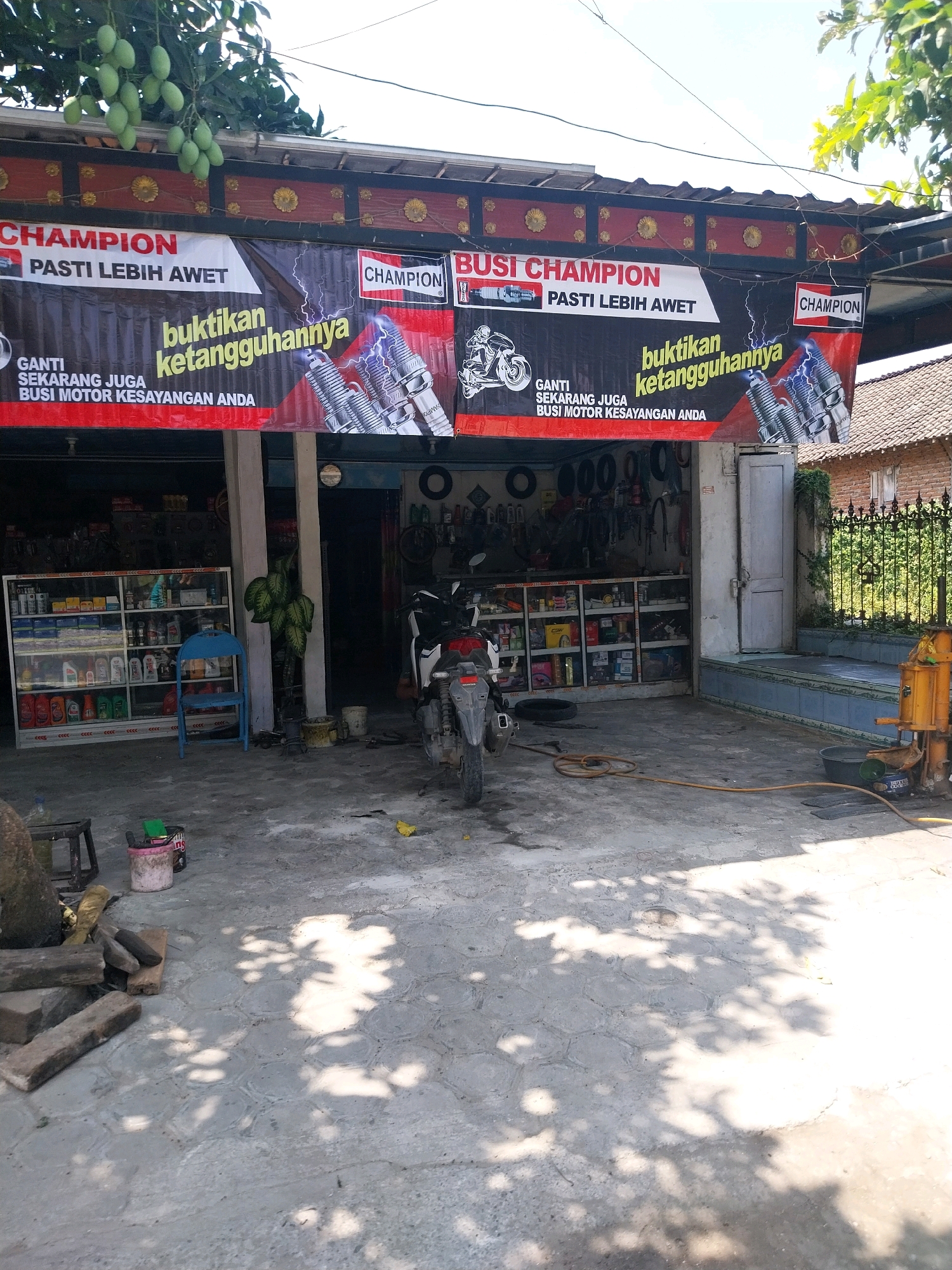 The image depicts the exterior of a motorcycle repair shop in Campur, Indonesia. A banner prominently displays the brand "CHAMPION" with the Indonesian slogan "PASTI LEBIH AWET" (Certainly More Durable) and "BUKTIKAN KETANGGUHANNYA" (Prove Its Toughness). The banner also shows images of spark plugs and a motorcycle. Inside the shop, shelves are stocked with various automotive fluids and parts. A blue plastic chair and a potted plant are placed outside the shop entrance. A motorcycle is parked near the shop, and a yellow air compressor is visible on the right side of the image. The ground is paved with concrete, and shadows of trees are cast across it. The overall scene suggests a commercial establishment focused on motorcycle maintenance and parts sales.