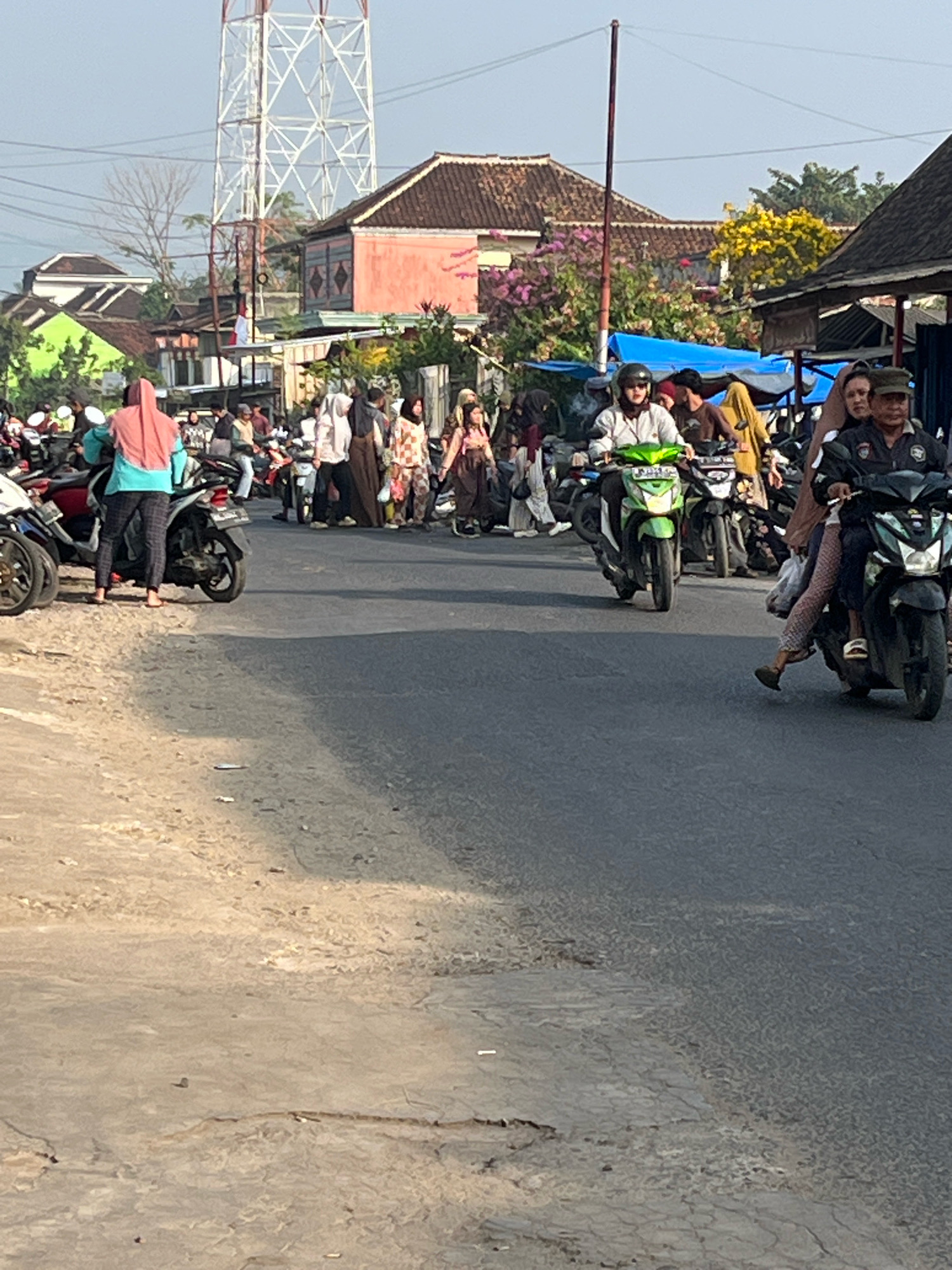 The image captures a street scene in Kemuning, Indonesia, likely during daylight hours given the bright illumination and shadows. The foreground is dominated by a paved road, with a textured, sandy shoulder on the left. Several motorcycles are parked and in motion on the road. In the middle ground, a group of people, predominantly women wearing headscarves, are gathered, some standing near motorcycles and others walking. One woman in a pink headscarf and teal top is standing on the shoulder, facing away from the camera.
Further back, more individuals are visible, with a mix of traditional and casual clothing. Buildings with tiled roofs line the street, some featuring colorful facades, including a prominent pink building. A tall, white communication tower stands in the background. Green and yellow foliage is present, suggesting a somewhat tropical environment. Two men on motorcycles are riding down the road; one is on a green scooter, and the other is on a black scooter. The overall impression is of a bustling street in a residential or commercial area, possibly a market or a gathering point.