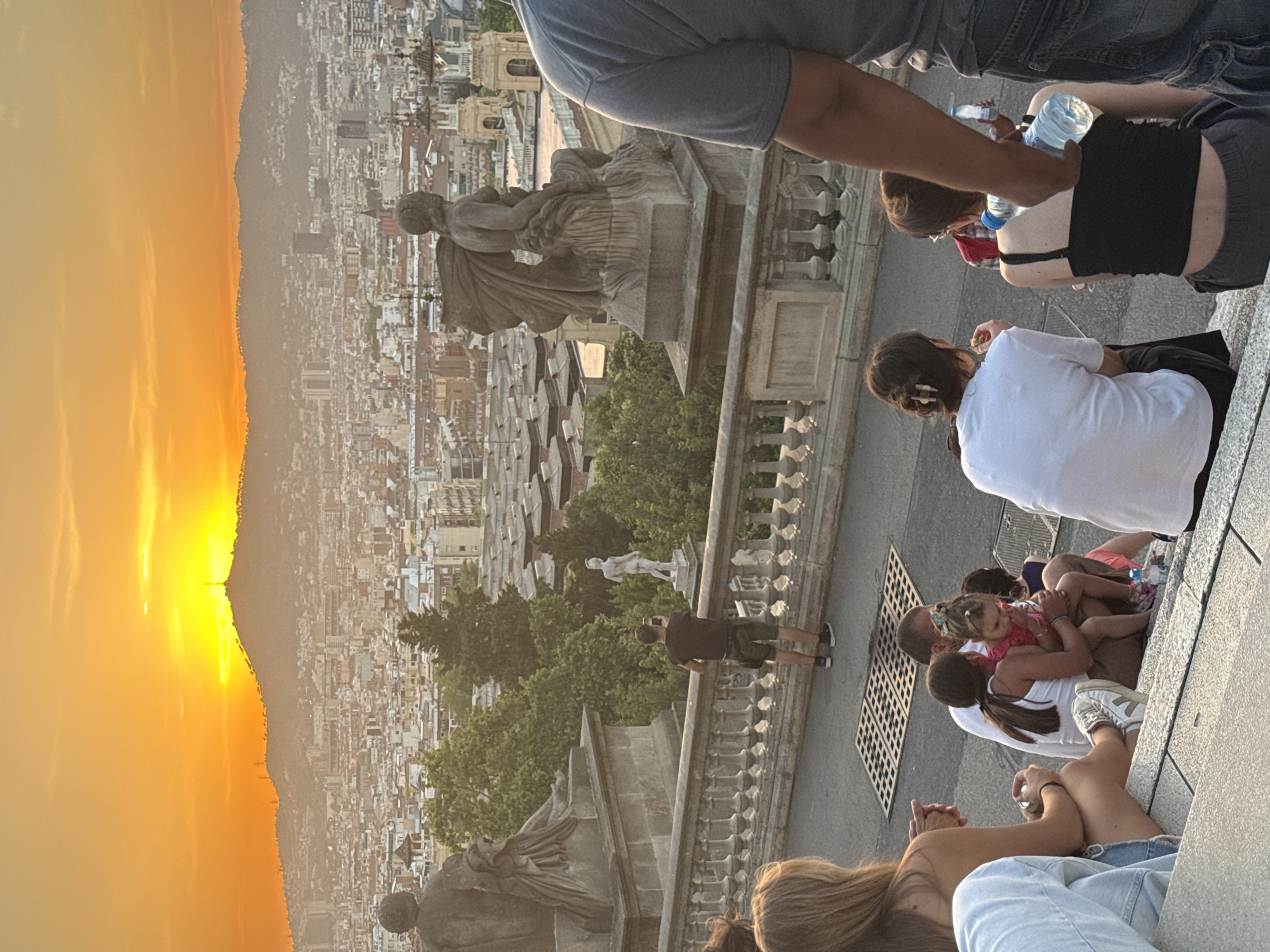 A wide shot captures a panoramic view of Barcelona, Spain, at sunset. The sky is ablaze with vibrant orange and yellow hues as the sun dips behind a distant, silhouetted mountain range that is topped with a radio tower. Below the mountain, the city sprawls out, a dense collection of buildings with terracotta roofs and lighter-colored structures.
In the foreground, a group of people are seated on wide stone steps. To the left, a person with long blonde hair sits facing away from the camera, wearing a light-colored top and denim shorts. Next to them, a family is gathered on the steps. A man in a white t-shirt sits with his arm around a young girl in a pink top. Another young child is partially visible behind them. A bottle of water is on the steps near them.
Further to the right, a person wearing a white t-shirt faces away from the camera, and to their right, a person in a black tank top and grey pants is seated. Their back is to the camera, and they are holding a bottle of water. Another person's arm, wearing a grey t-shirt, is in the far right of the frame.
Behind the seated individuals, a stone balustrade with decorative railings runs horizontally. Beyond the balustrade, two large, ornate statues are visible on pedestals. One on the left appears to be a seated figure. The other on the right is a seated female figure with wings. Between the statues and the city, lush green trees are interspersed with the architecture. A lone figure, a man in a dark t-shirt and shorts, stands near the balustrade, facing the city and holding up a smartphone, appearing to take a photograph. Another smaller statue is visible in the background amongst the trees. The scene is peaceful, with the city bathed in the warm glow of the setting sun.