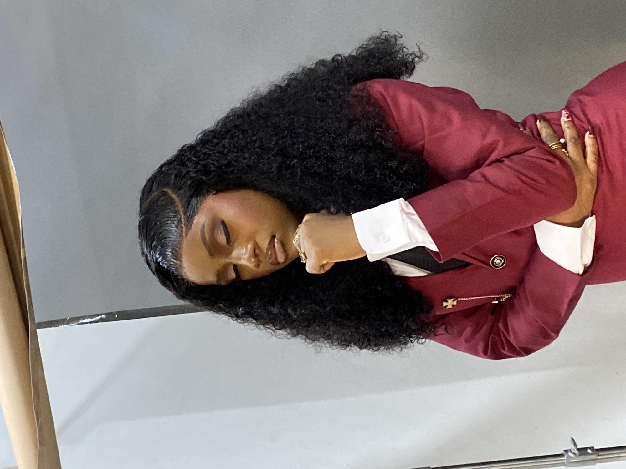 An adult female with dark skin and long, dark, curly hair, styled with laid edges and a left side part, is depicted from the waist up. Her eyes are closed, and her head is tilted slightly downward and to her right, with her right hand, lightly fisted and adorned with multiple gold-toned rings, resting against her chin. Her left hand, also bearing rings and dark manicured nails, is partially visible at her waist. She is wearing a white collared shirt, a dark tie, and a maroon/burgundy blazer featuring gold-toned buttons. A gold-toned brooch with a cross design and a chain is pinned to the left lapel of her blazer. The background consists of plain light grey and off-white walls, indicative of an indoor studio setting. A metal stand or tripod is visible on the far left, and a brown rolled material is present at the top left corner of the frame. The specific city location of Gura Top, Nigeria, cannot be determined from the visual information presented in the image.