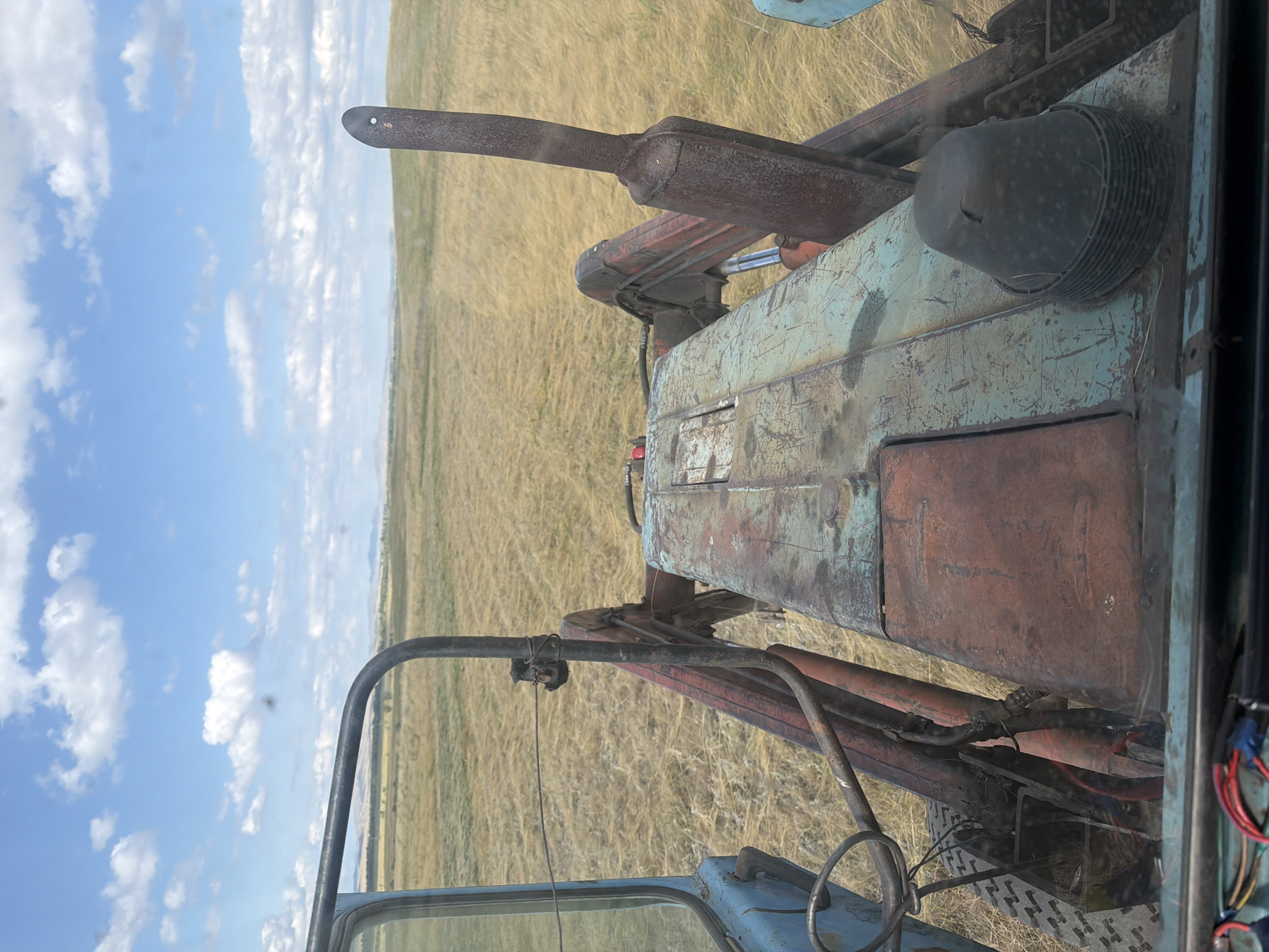The image depicts a view from the driver's seat of a blue tractor, possibly in the village of Khalilovo, Russia. The tractor's weathered dashboard and controls are visible in the foreground, with a rusty front loader mechanism dominating the center of the frame. Beyond the tractor, a vast, dry field of golden grass stretches towards a hazy horizon under a blue sky dotted with white clouds. The scene suggests agricultural activity or the aftermath of harvesting. No people are visible in the frame.
