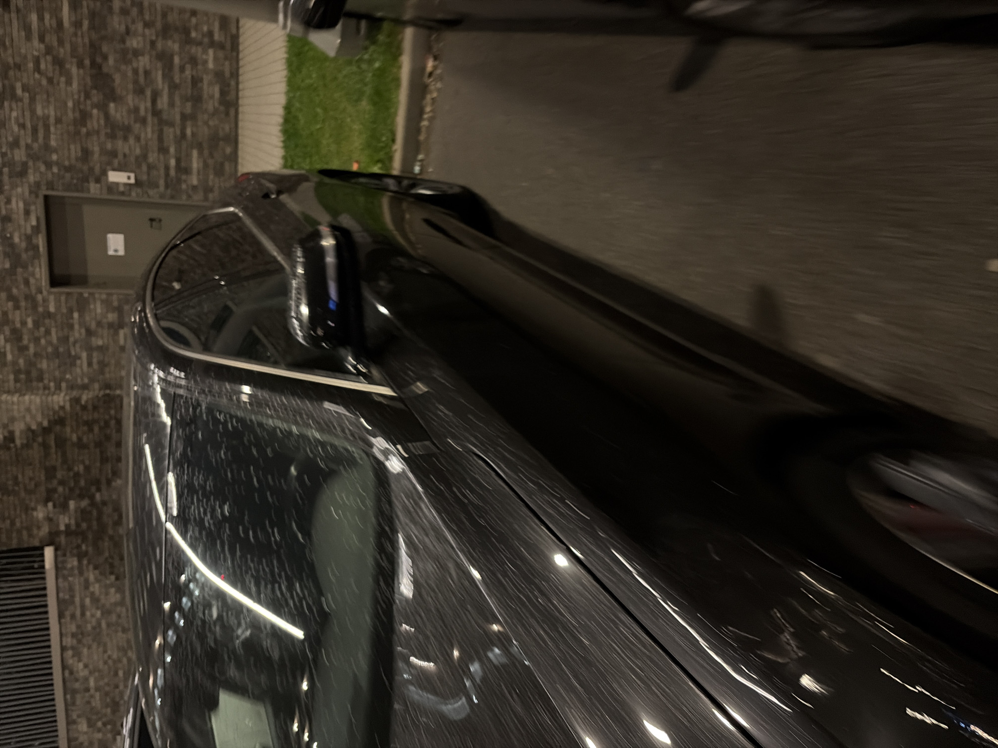 A dark sedan is parked on asphalt, with a grass verge and a brick building in the background. The car's surface is wet, with numerous reflections and streaks of light, suggesting it may be raining or has recently rained. A portion of another vehicle is visible on the right side of the frame. The building features a door with a light fixture and a sign. The scene appears to be an outdoor parking area.