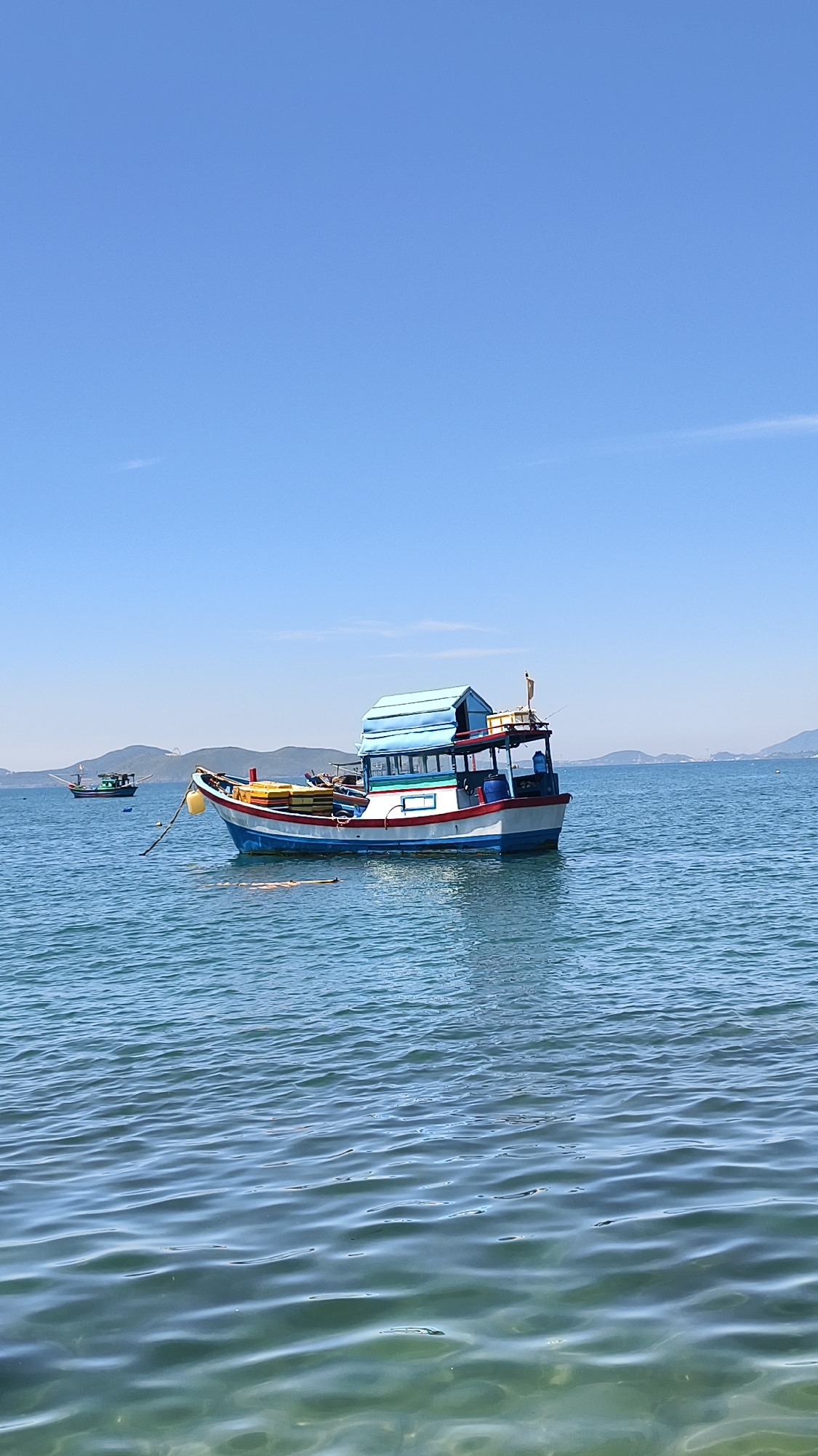 In Bac Nha Trang Ward, Vietnam, a clear blue sky stretches over the tranquil sea. Two fishing boats are anchored in the water. The larger boat, in the foreground, is painted white and blue with red trim, featuring a light blue roof structure. Yellow and green floats are visible on its deck, and it is tethered by a rope. A smaller, darker boat is anchored further in the distance. The water's surface is calm with gentle ripples, and distant, hazy hills can be seen on the horizon. The scene is brightly lit by the sun.